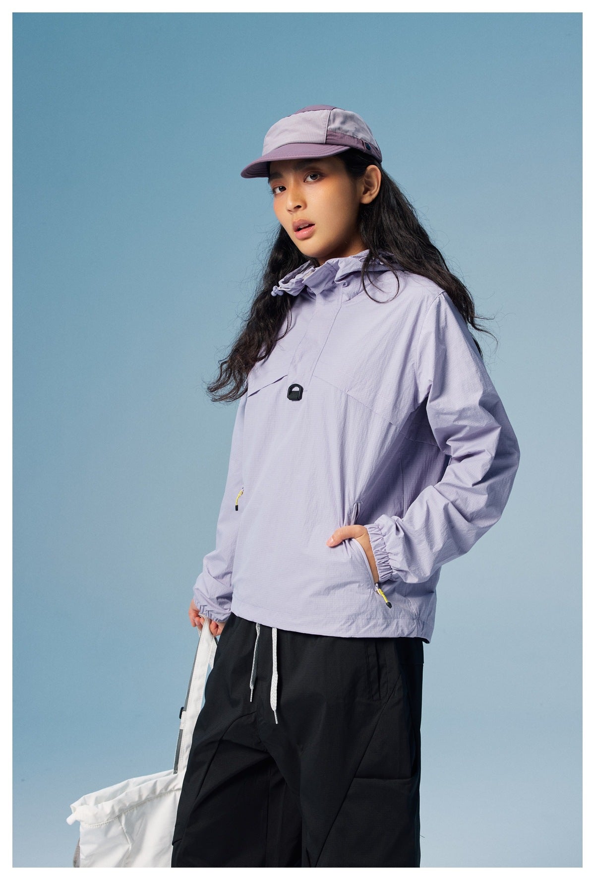 Model in a light lavender hooded pullover, black pants, and a matching cap, holding a white bag.