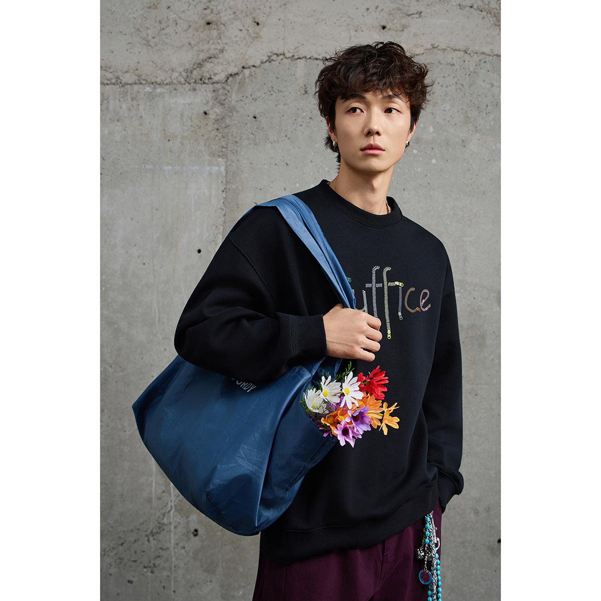 Model wearing a black crewneck sweatshirt with colorful lettering and a flower graphic, carrying a large blue tote.