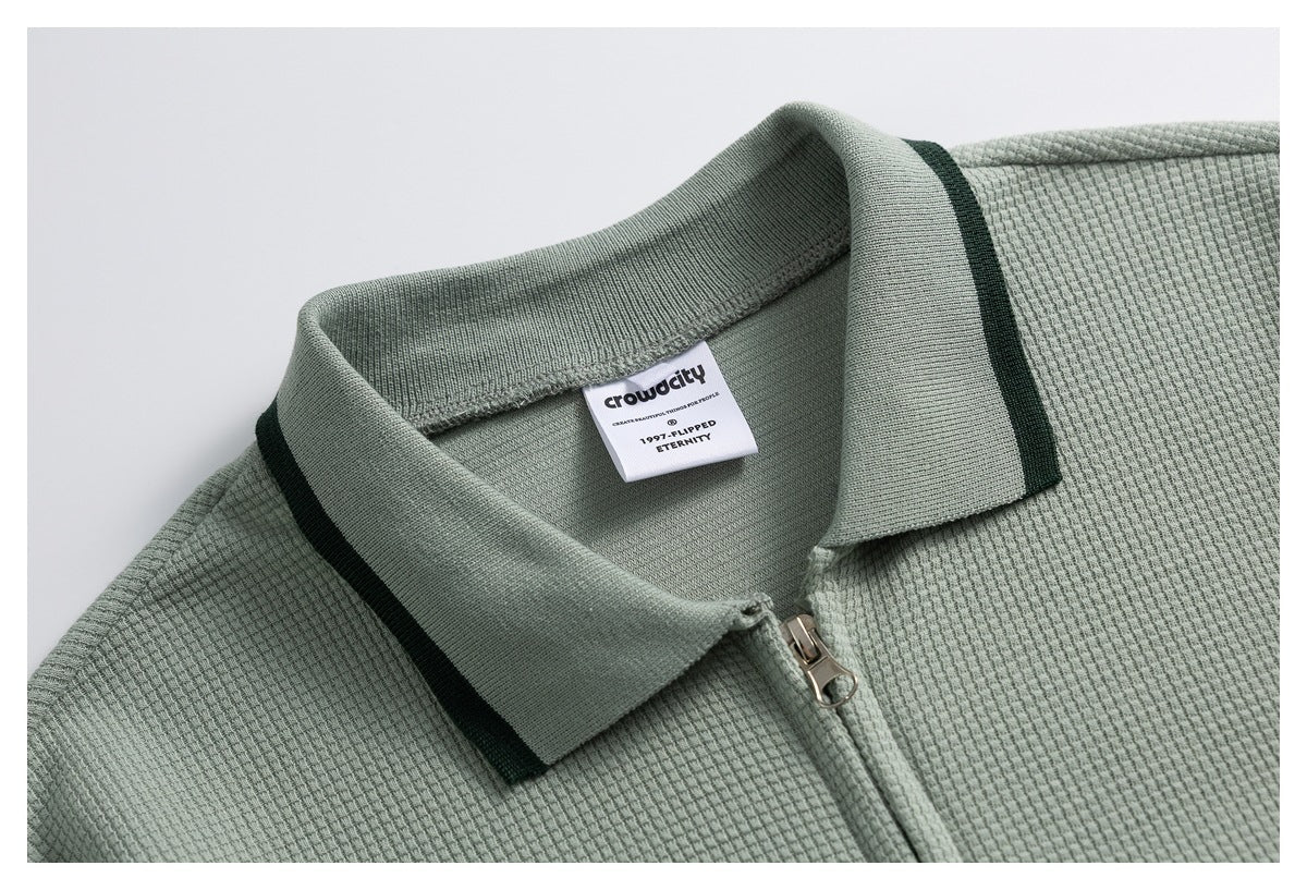 Close-up of a sage green textured zip-up top with a pointed collar, dark green trim and a visible neck label.