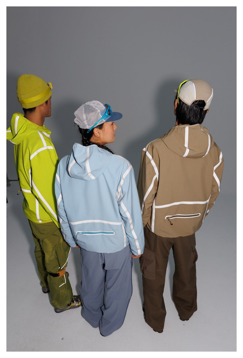Rear view of three models wearing neon yellow-green, pale blue, and tan hooded jackets with contrasting trim.