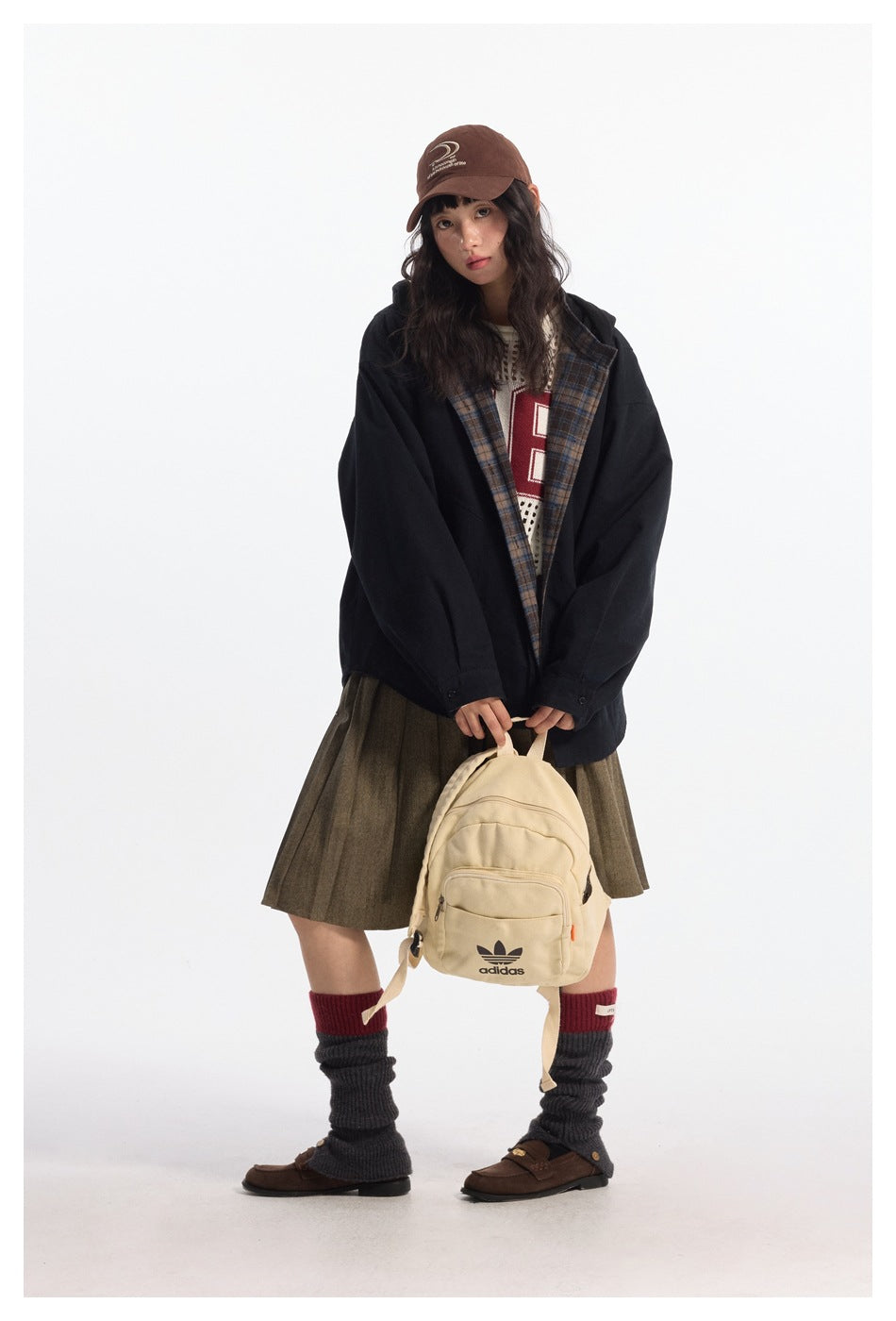 Model wearing a black jacket with plaid lining and an olive pleated skirt, holding a beige backpack.
