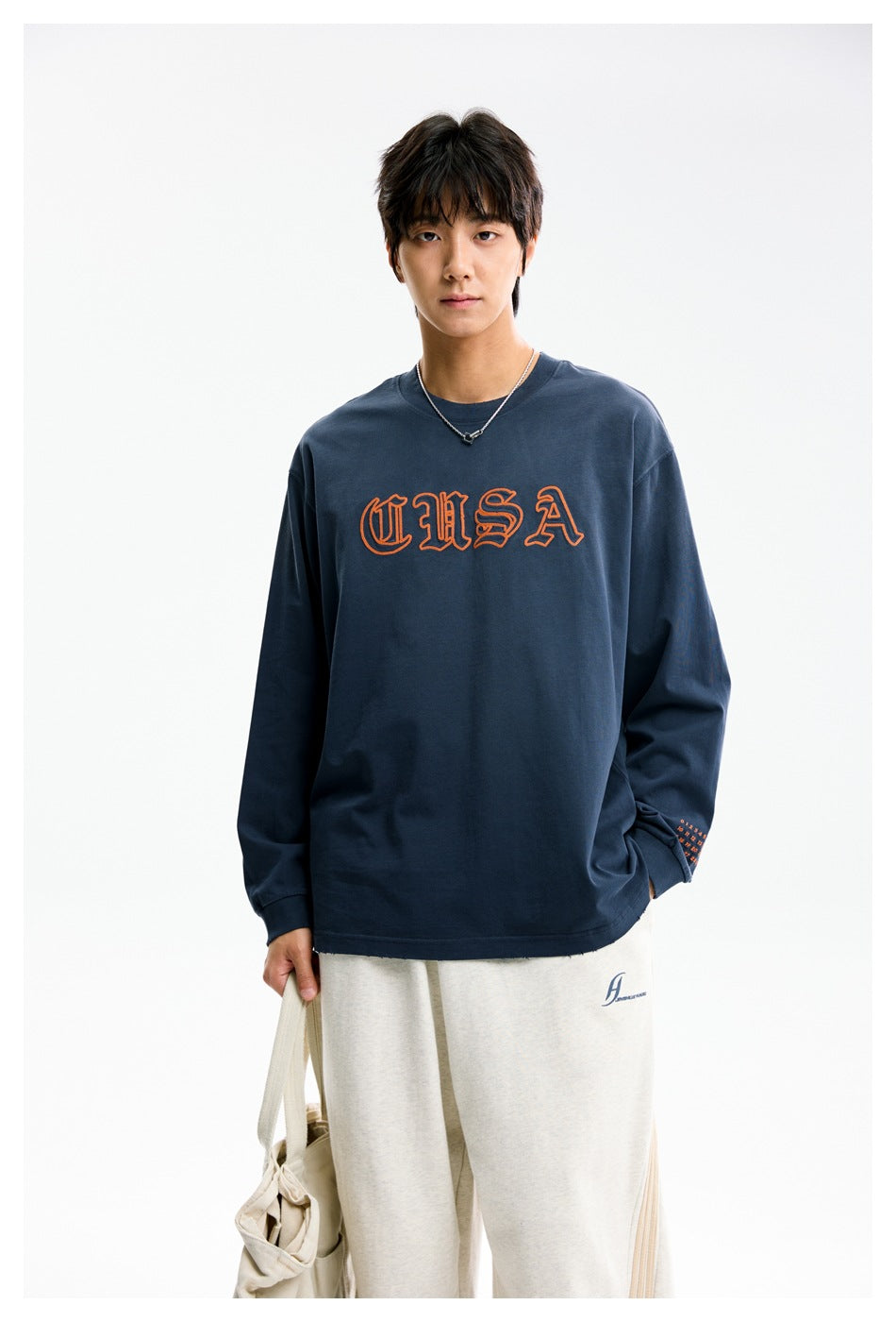 Model wearing a navy long-sleeve shirt with orange outlined lettering and light-colored pants.