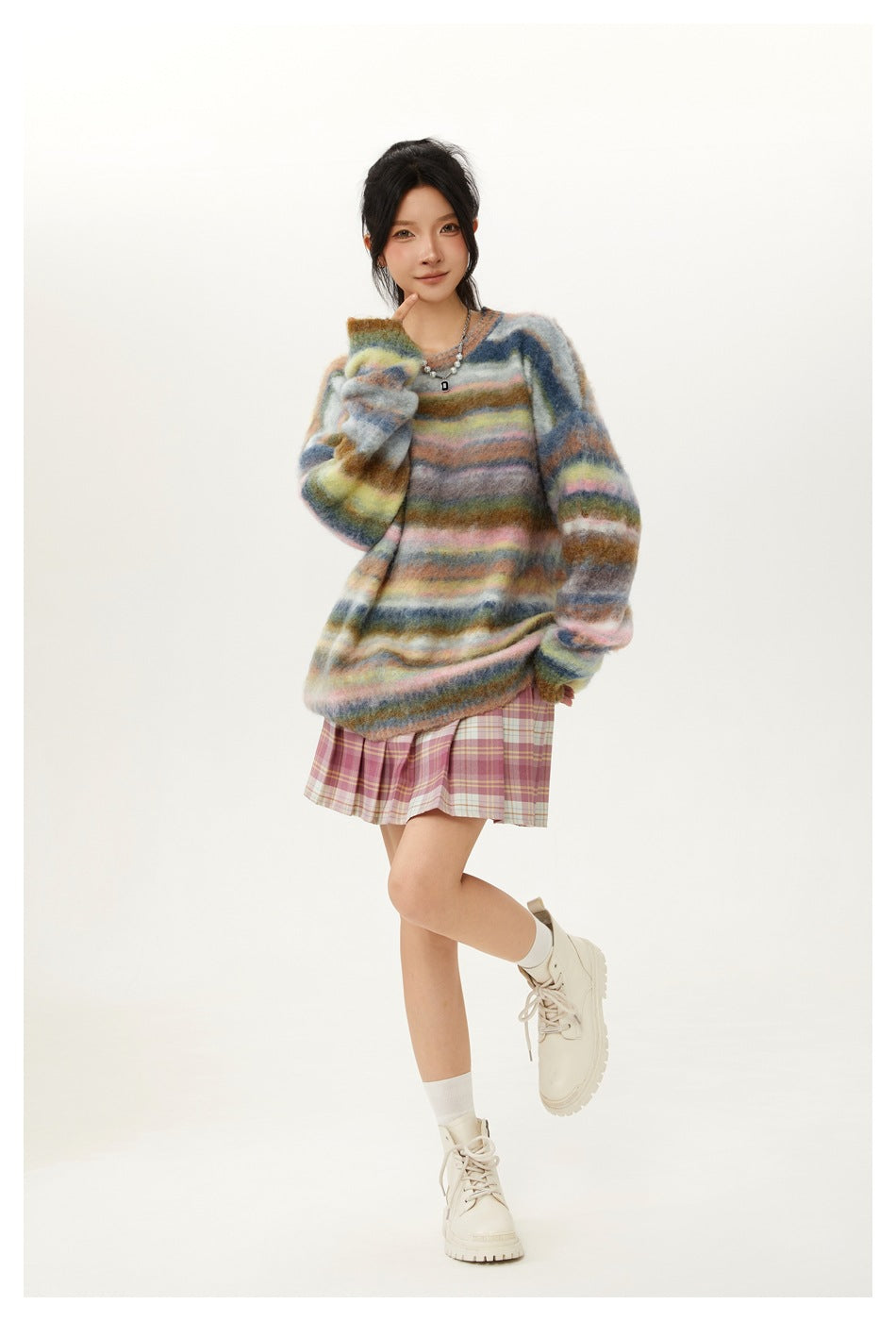 Model posing in a loose multicolor striped knit sweater with a pink plaid pleated skirt and cream boots