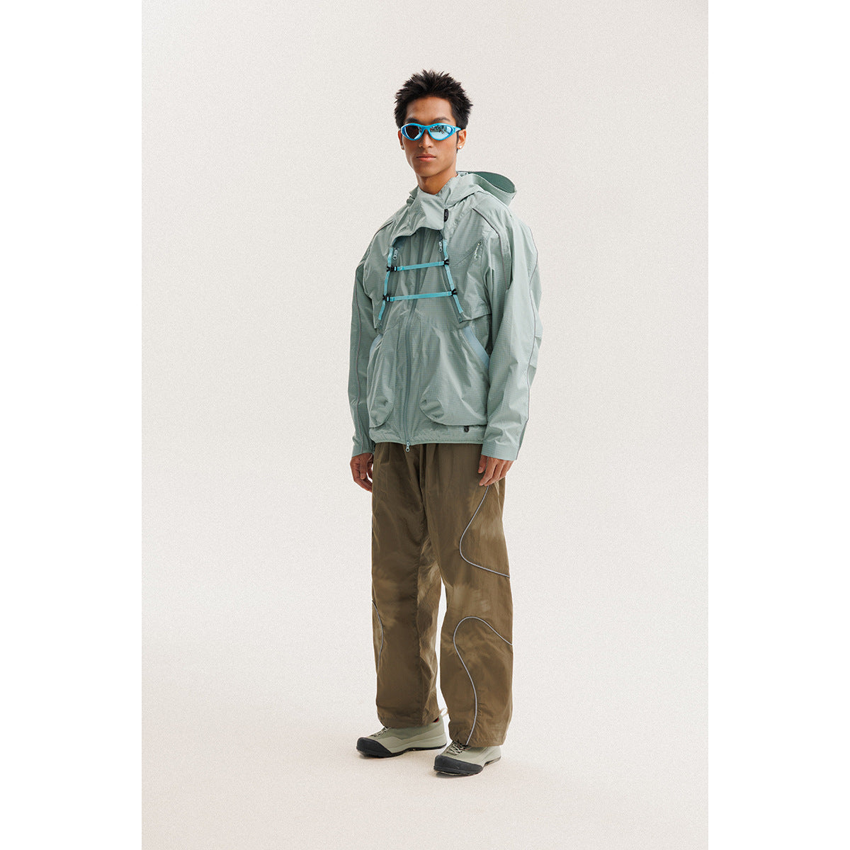 Model wearing a pale sage hooded jacket with blue chest straps and loose brown pants.