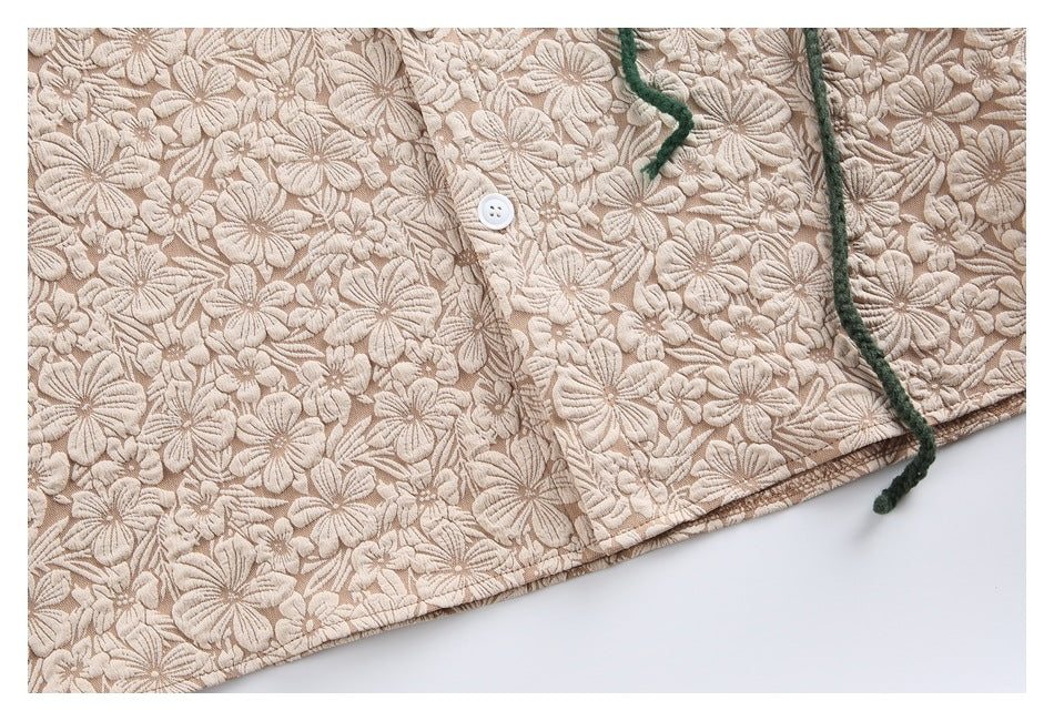 Close-up of the beige floral-textured garment's lower hem, white button placket, and a dangling green cord.