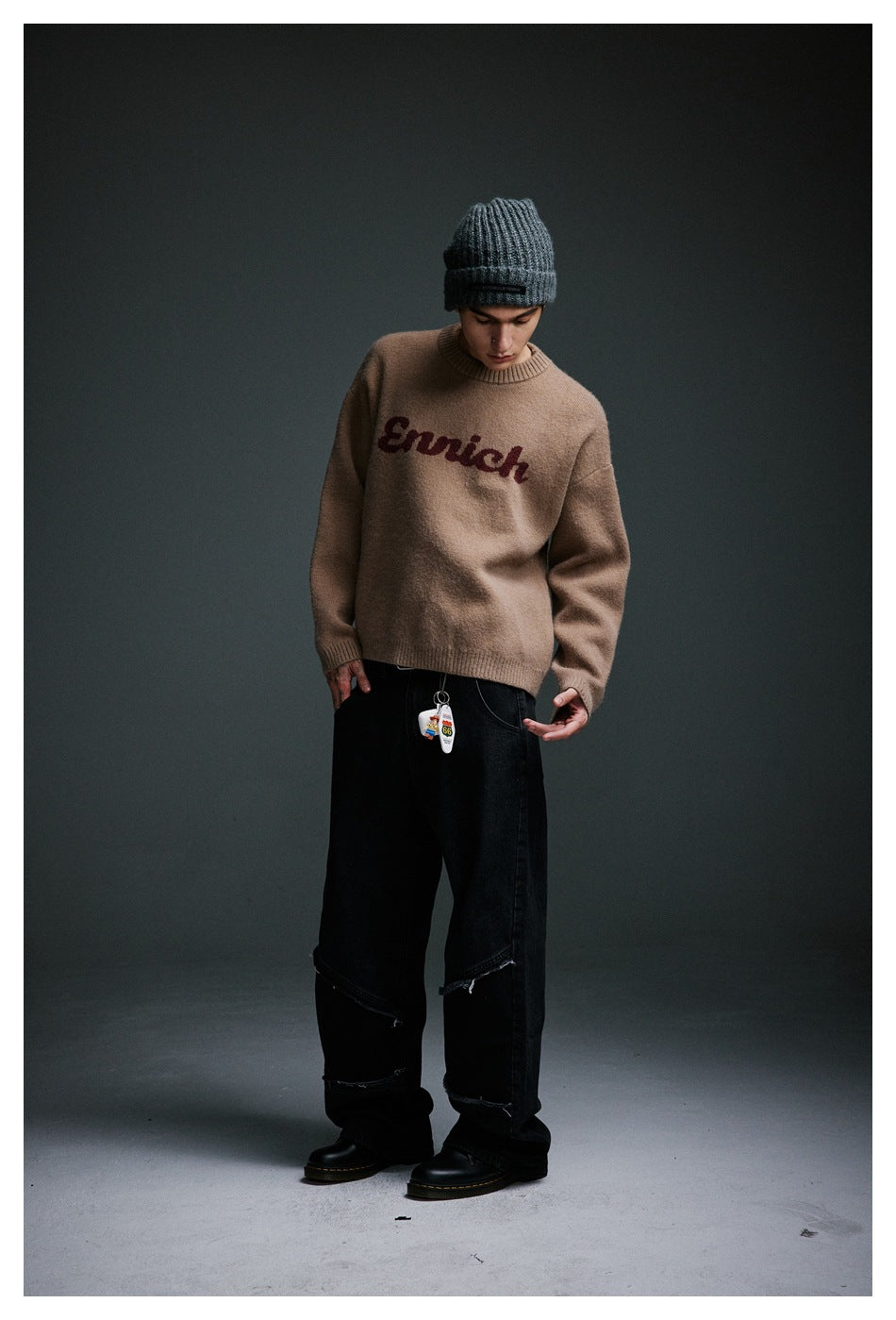 Full-length model wearing a taupe crewneck sweater with burgundy lettering, black jeans, and a charcoal knit beanie.