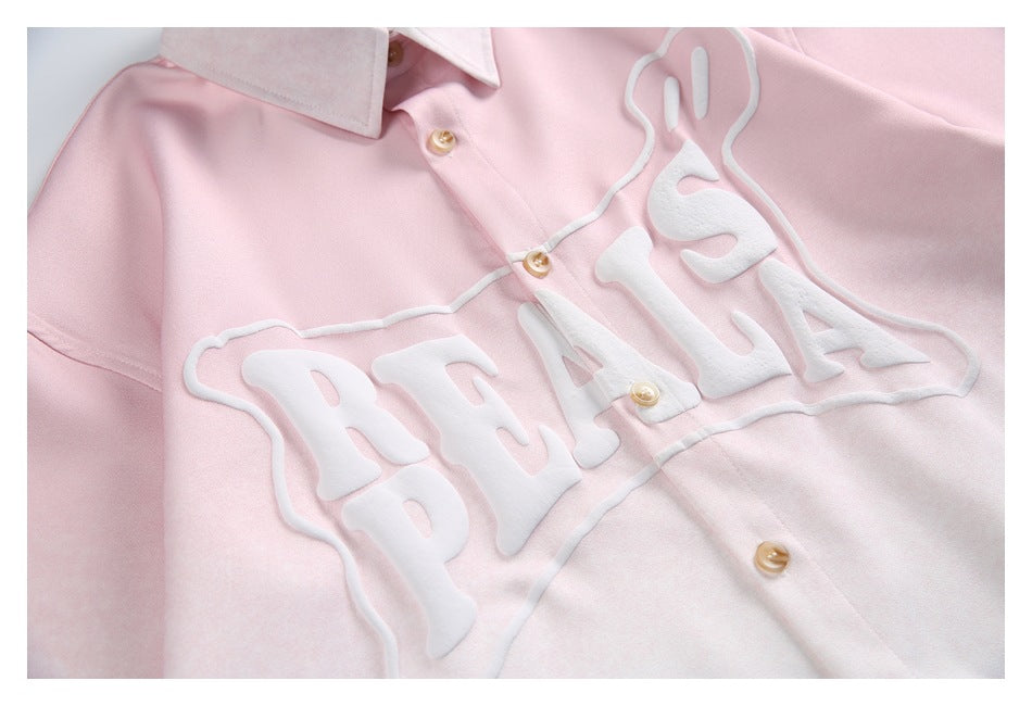 Close-up of a pale pink button-up shirt with a white collar, tan buttons, and raised white lettering on the chest.