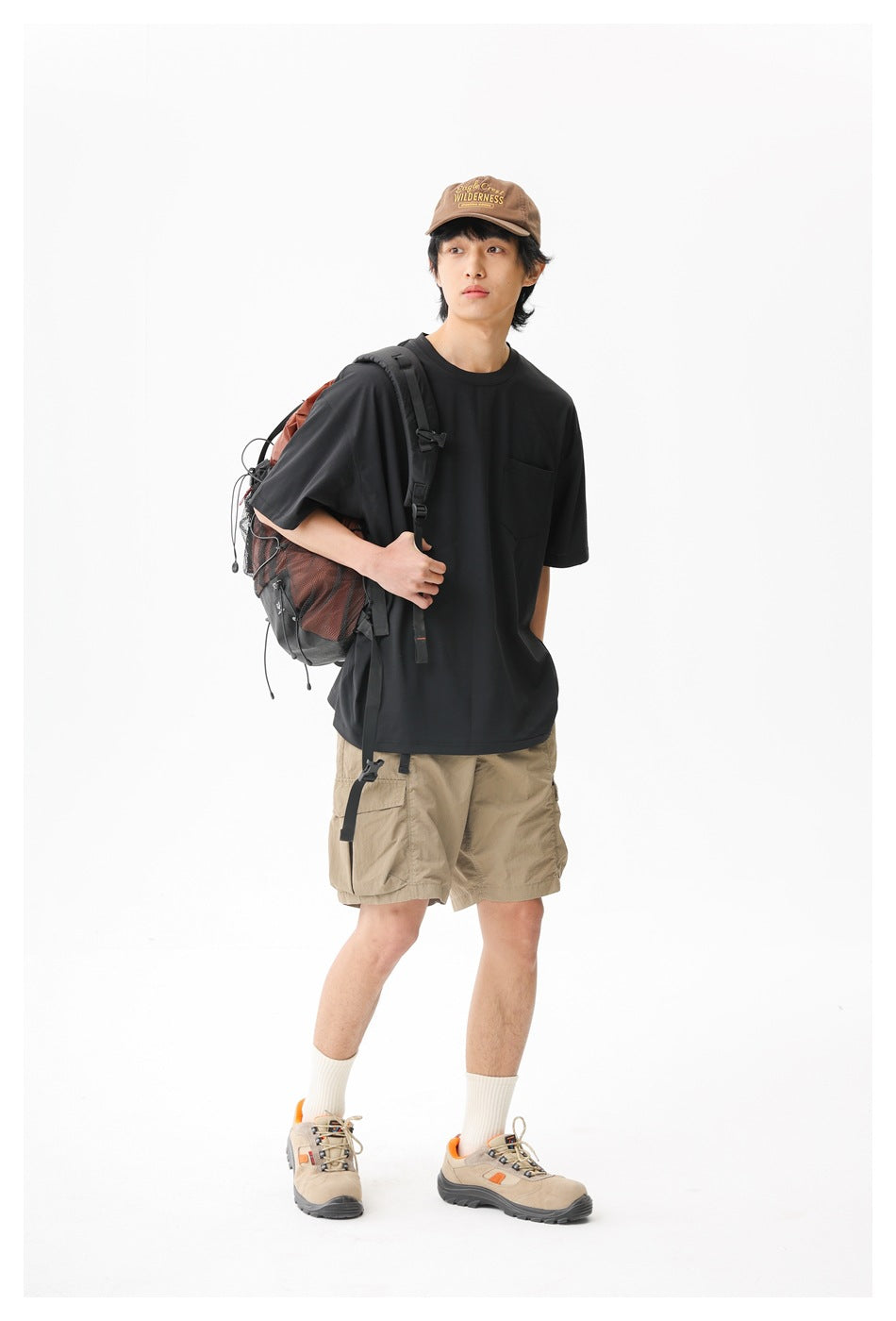 Model wearing a black pocket T-shirt and tan cargo shorts, carrying a hiking backpack.