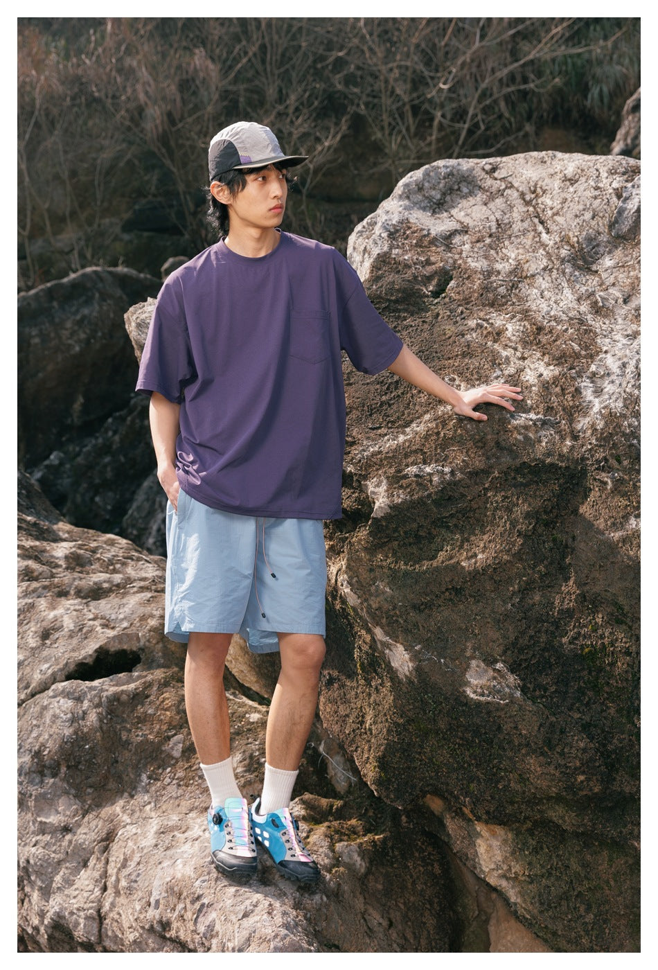 Model wearing a purple pocket T-shirt and light blue shorts, standing beside a large rock.