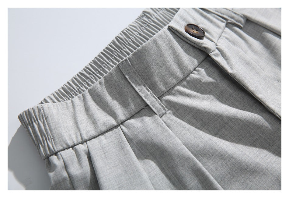 Close-up of light gray trousers showing the elasticized waistband, belt loop, button, and front pleat.