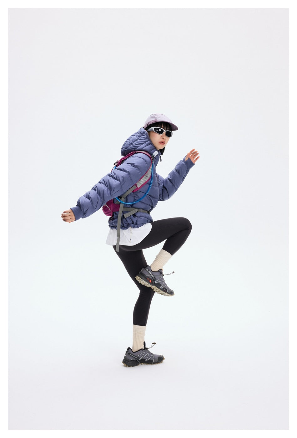 A model poses in a blue-purple hooded puffer jacket, black leggings and a backpack, lifting one knee.