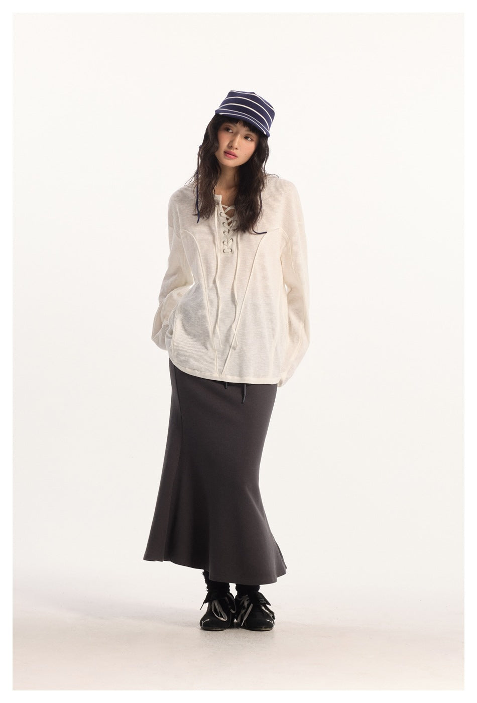 Full-length model wearing an ivory oversized lace-up top, dark flared maxi skirt, and navy striped cap.