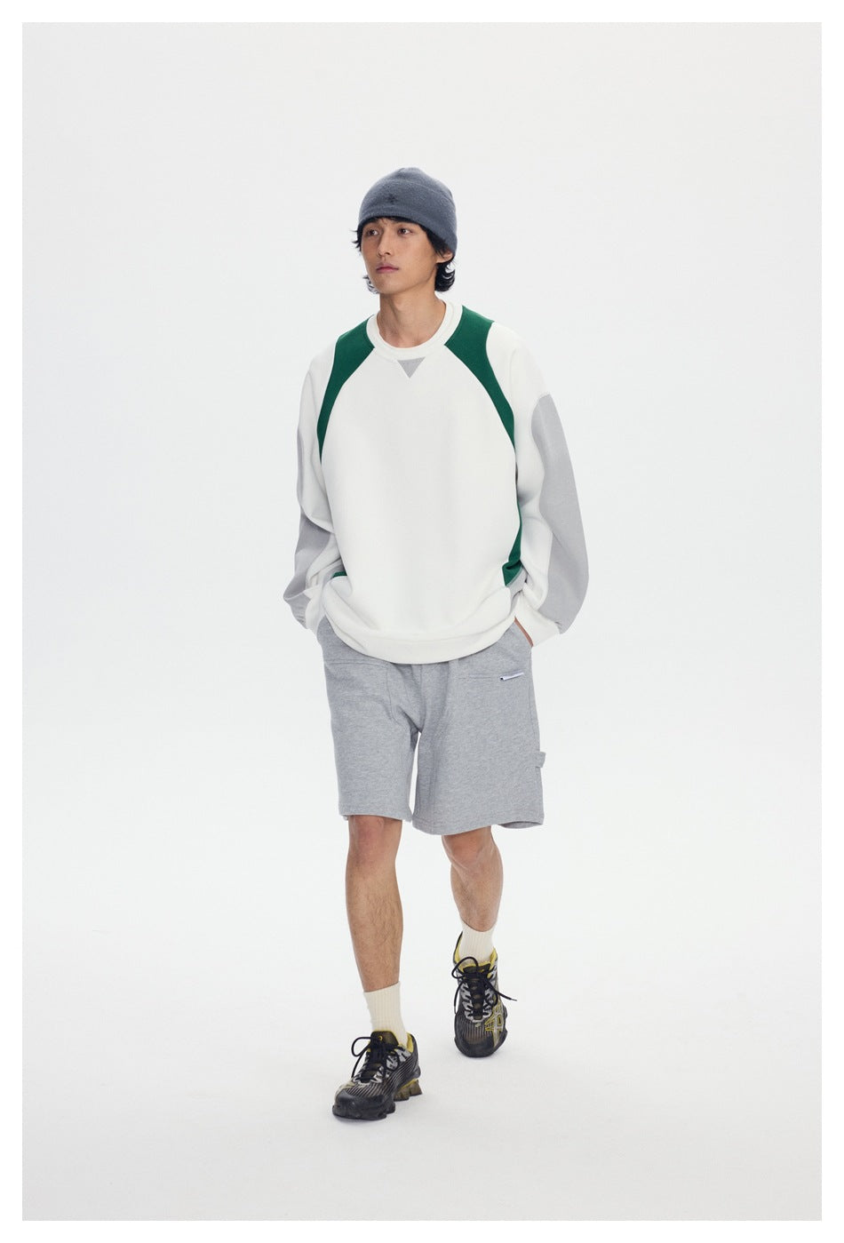 Model walking toward the camera in a white sweatshirt with green and gray panels and gray shorts.
