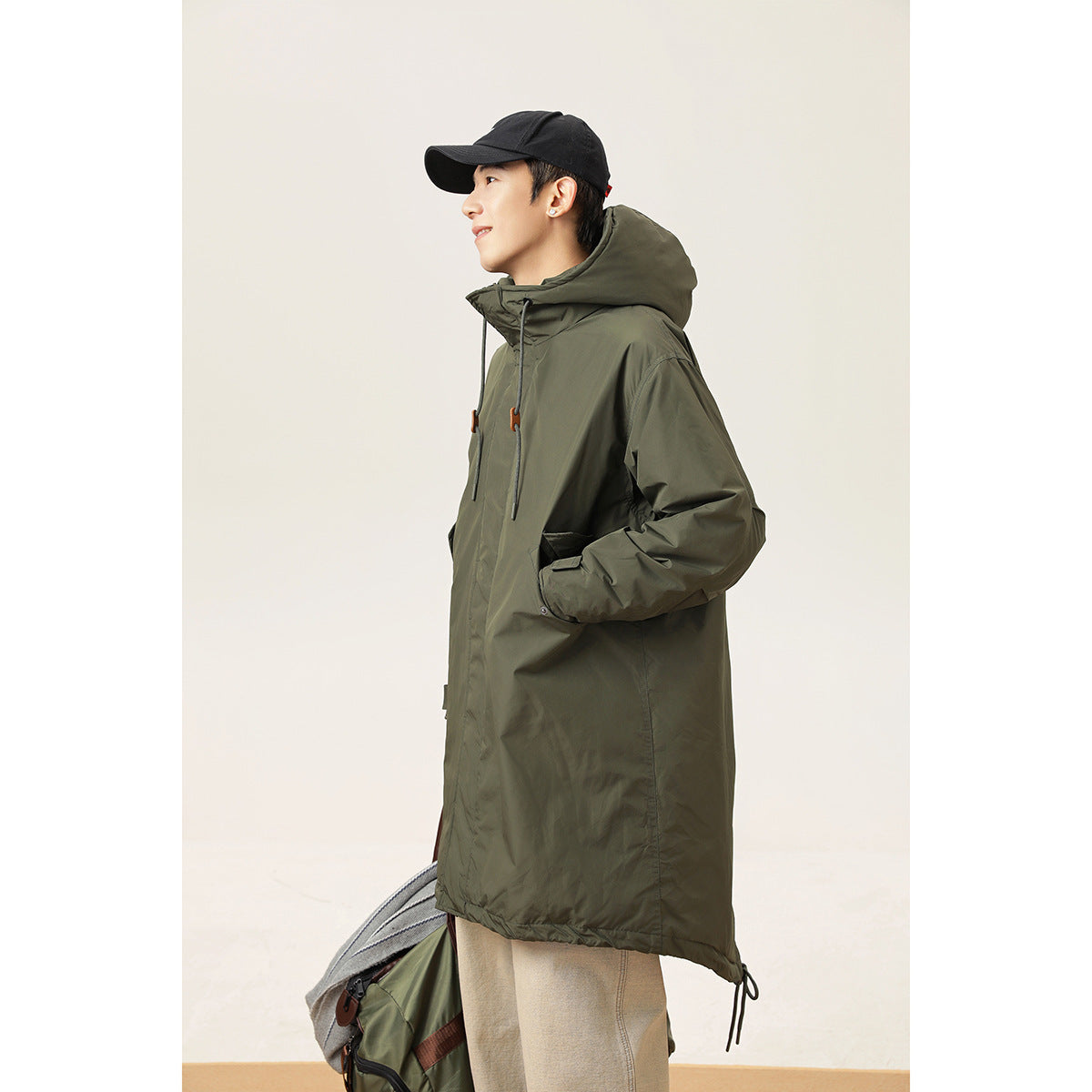 Side view of a model wearing a long dark olive hooded parka with front pockets and orange cord toggles.
