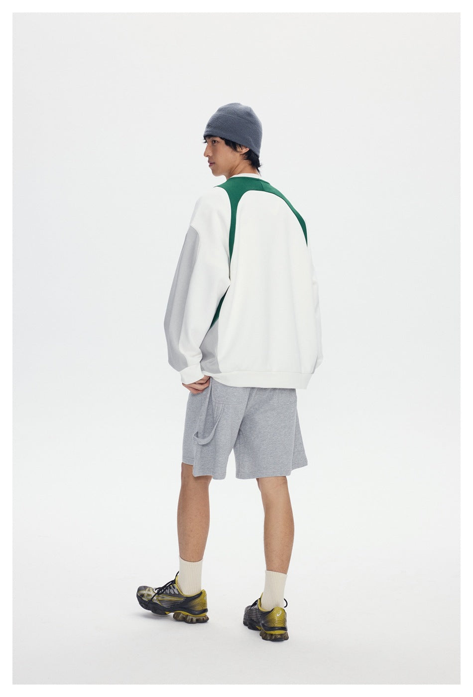 Model in gray knee-length shorts, back view, with a side loop detail, white and green sweatshirt and beanie