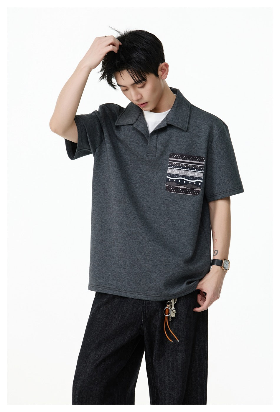 Model wearing a charcoal short-sleeve polo with a patterned chest pocket, shown in a relaxed pose.