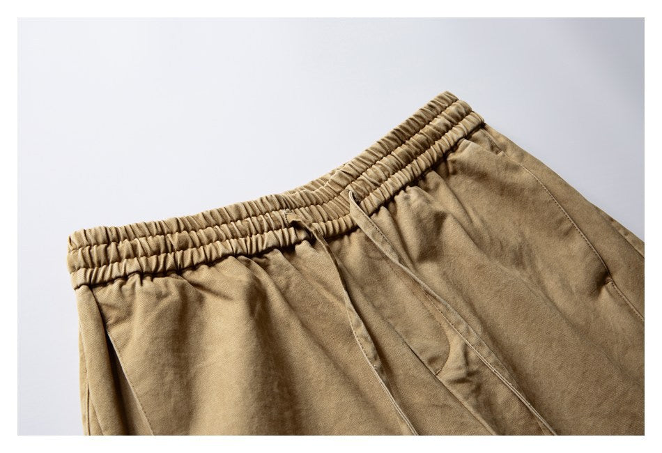 Close-up of tan trousers showing the gathered elastic waistband, drawstring and angled front pocket.