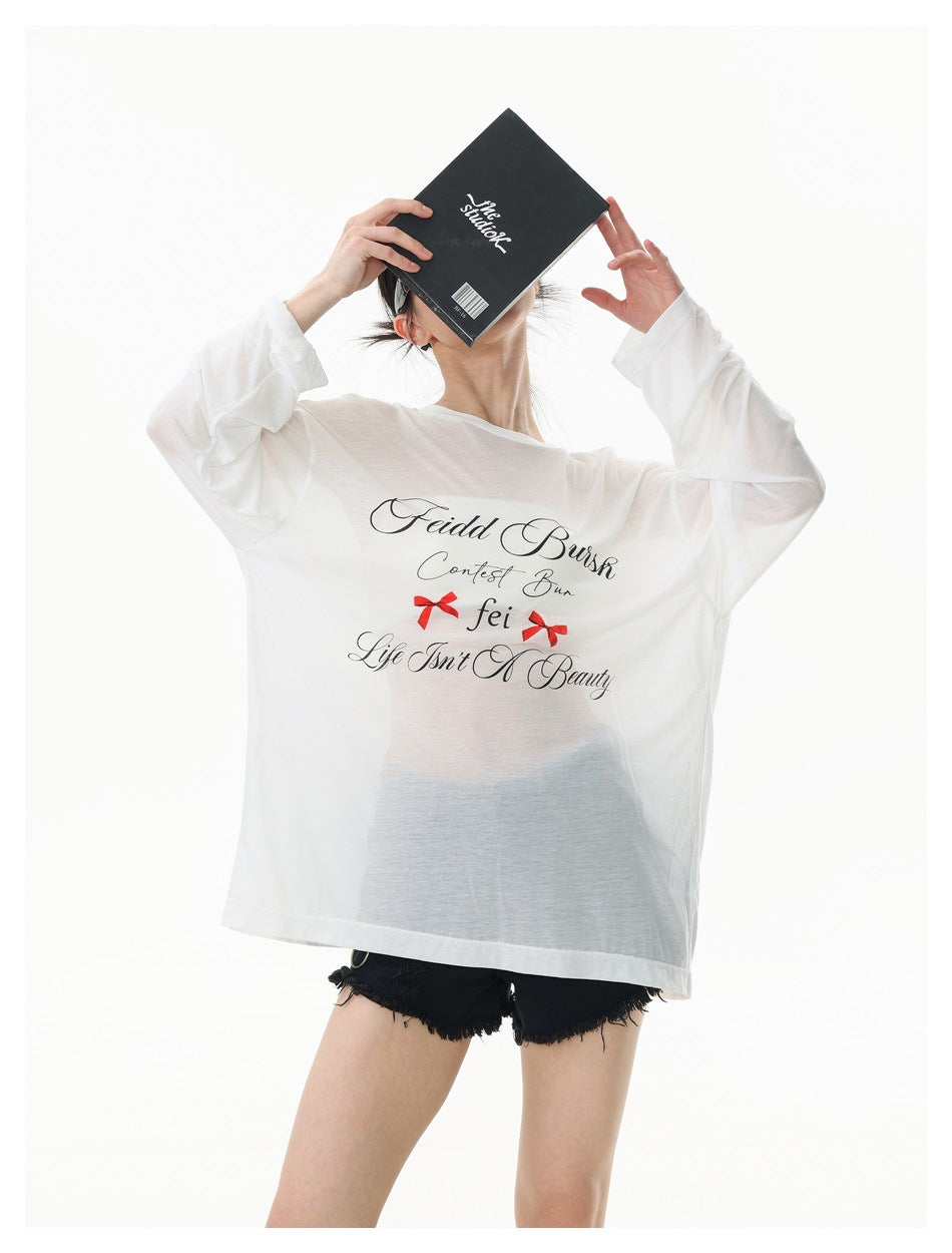 Model in a semi-sheer white long-sleeve top with script print and red bows, holding a book over her face
