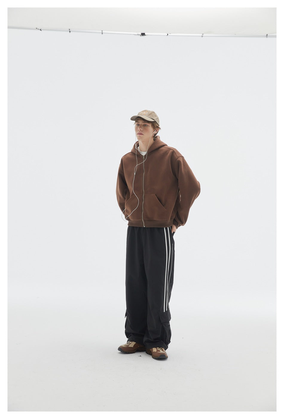 Model with hands in pockets wearing a brown zip-up hoodie and black striped cargo pants