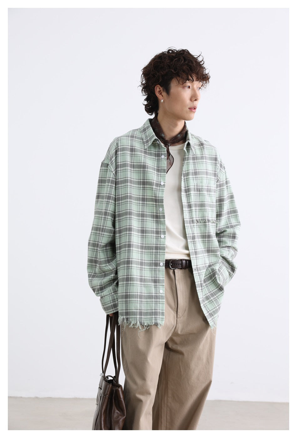 Model wearing an open green plaid shirt with frayed hem over a white ribbed tank, with a brown scarf and khaki pants