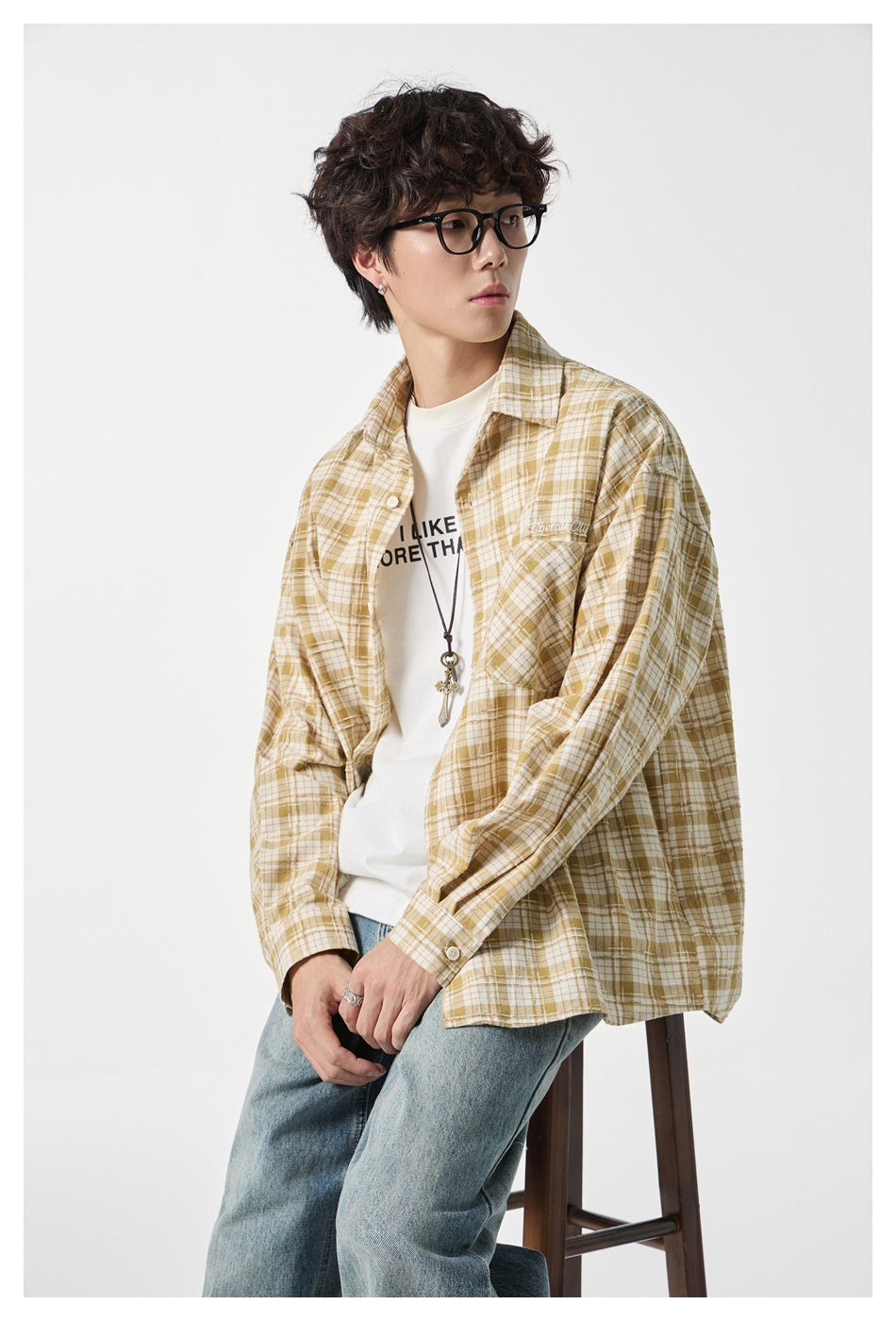 Seated model wearing a mustard and cream plaid button-up shirt over a white tee and loose blue jeans.