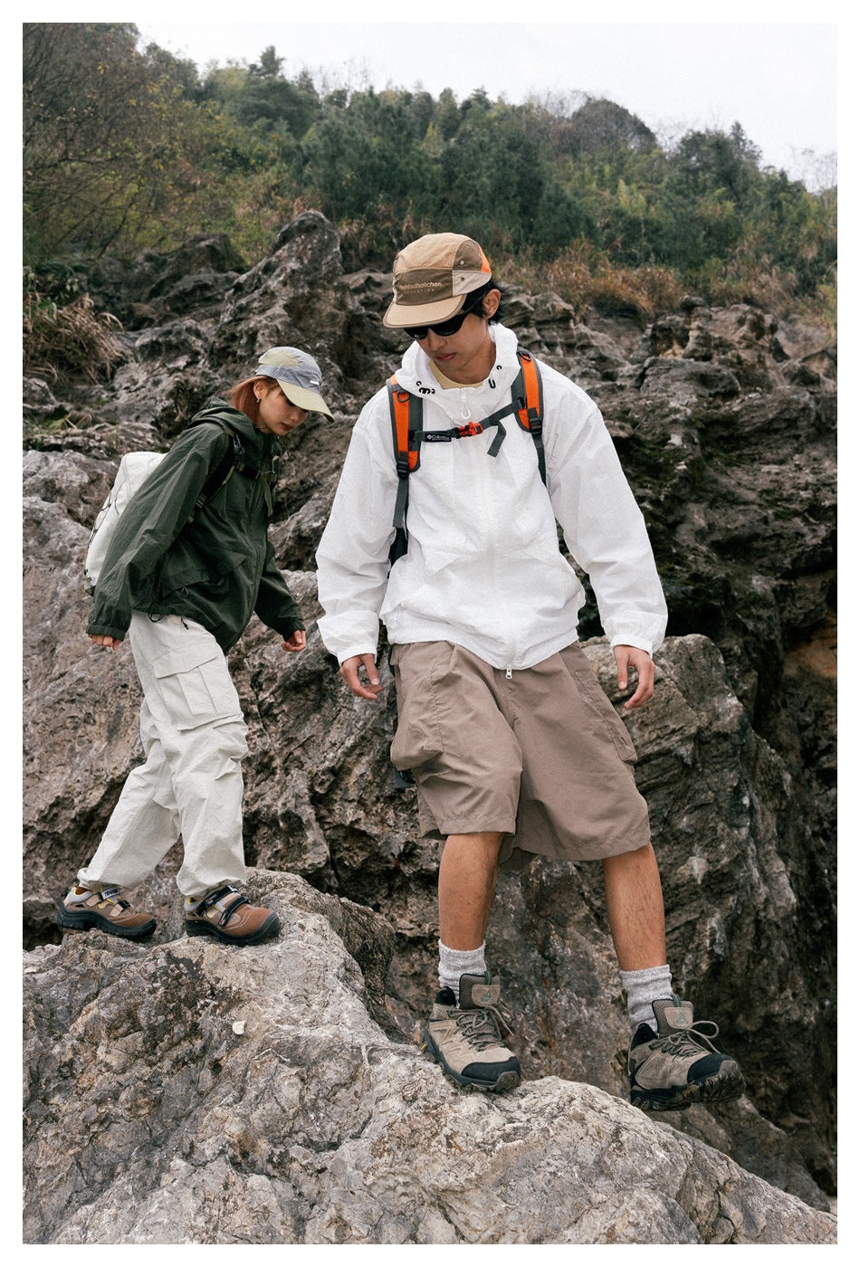 Two hikers on a rocky trail; the man wears a white hooded jacket and taupe shorts, and the woman a dark jacket.