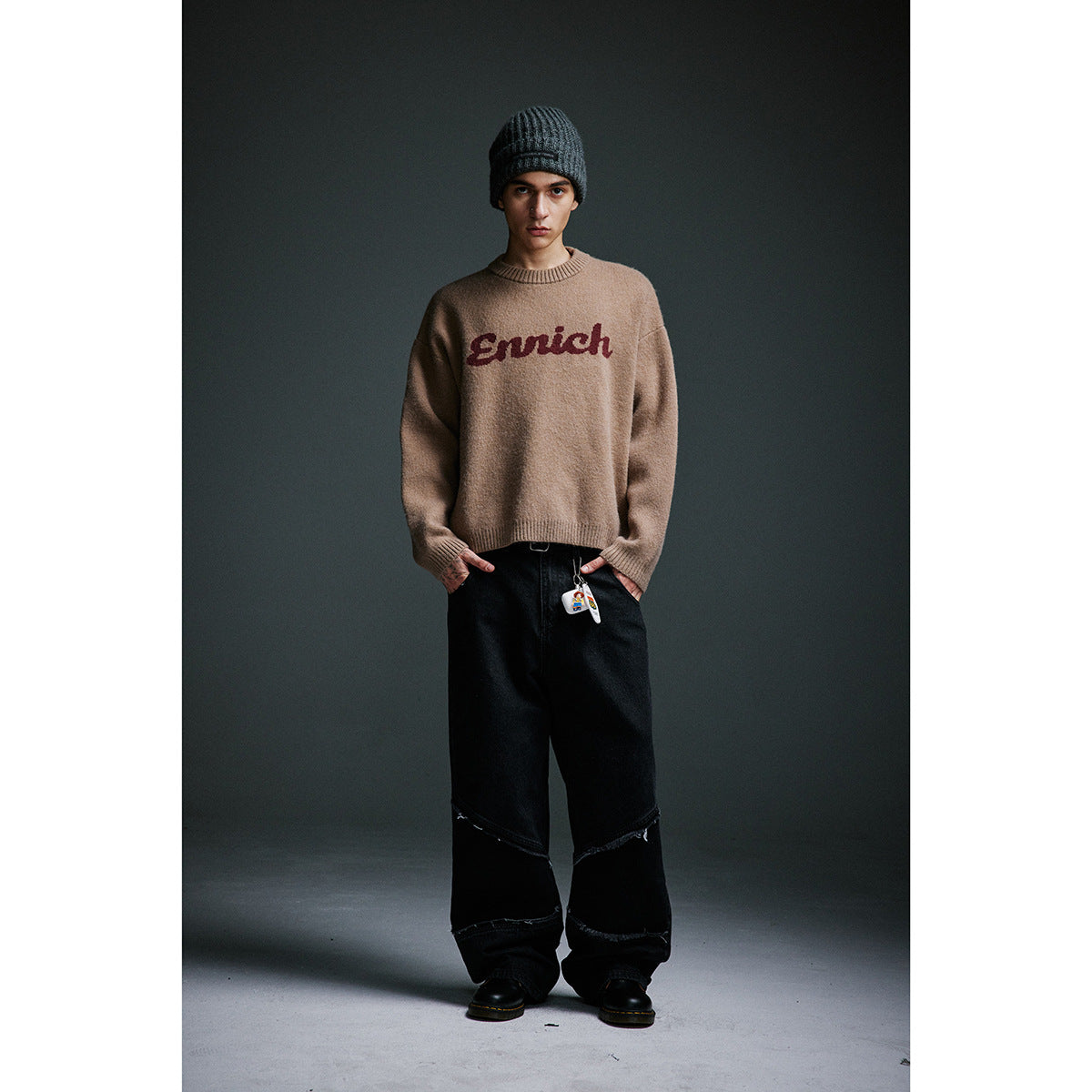 Full-length model wearing a tan sweater with burgundy script lettering, loose black jeans, and a dark ribbed beanie.