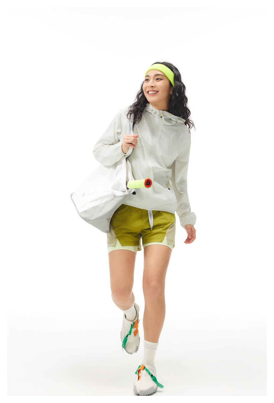 Model wearing an off-white hooded jacket and layered olive shorts, carrying a large white tote and a racket.