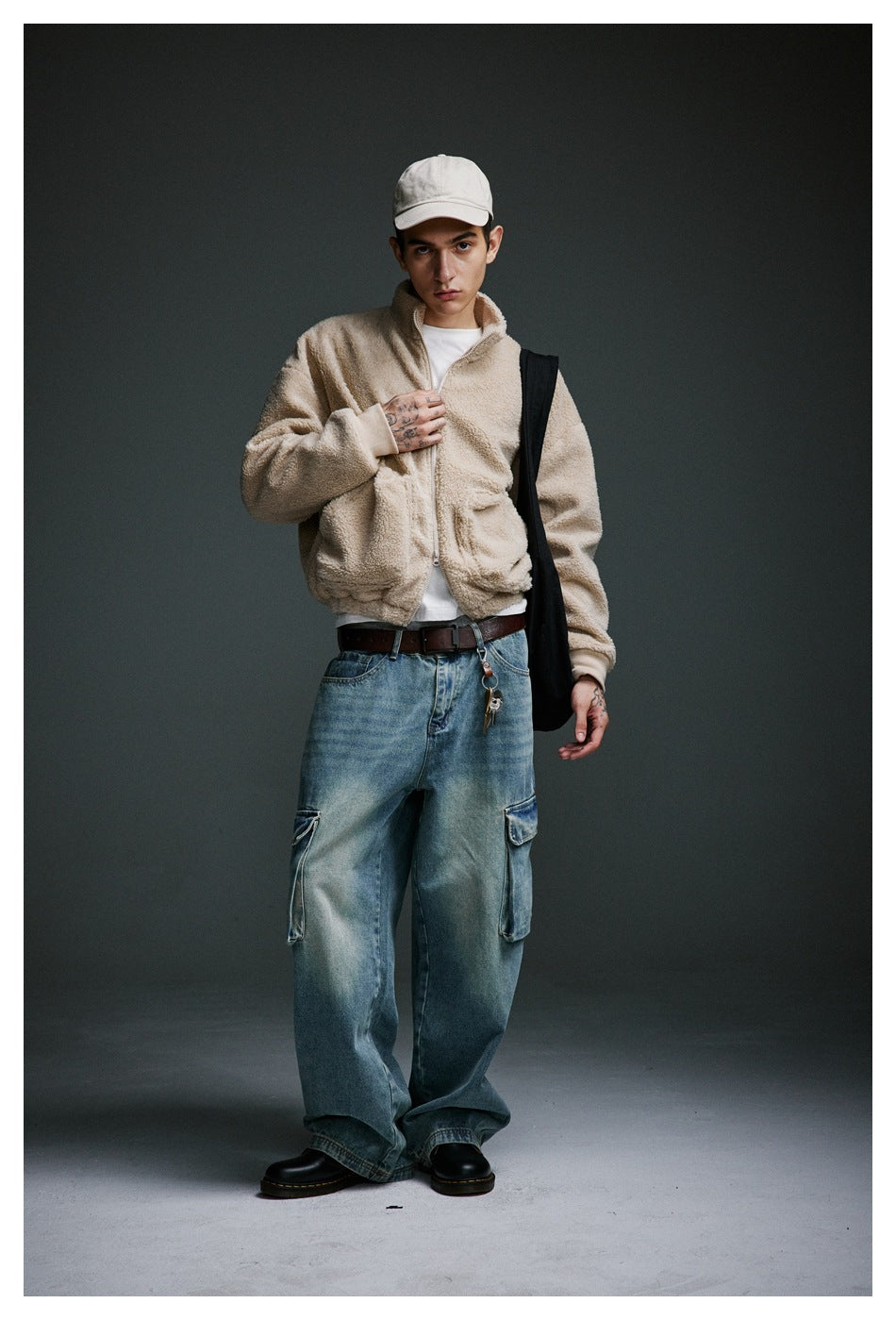 Full-length model wearing a cream zip-up jacket with oversized blue cargo jeans, white cap and black shoulder bag.