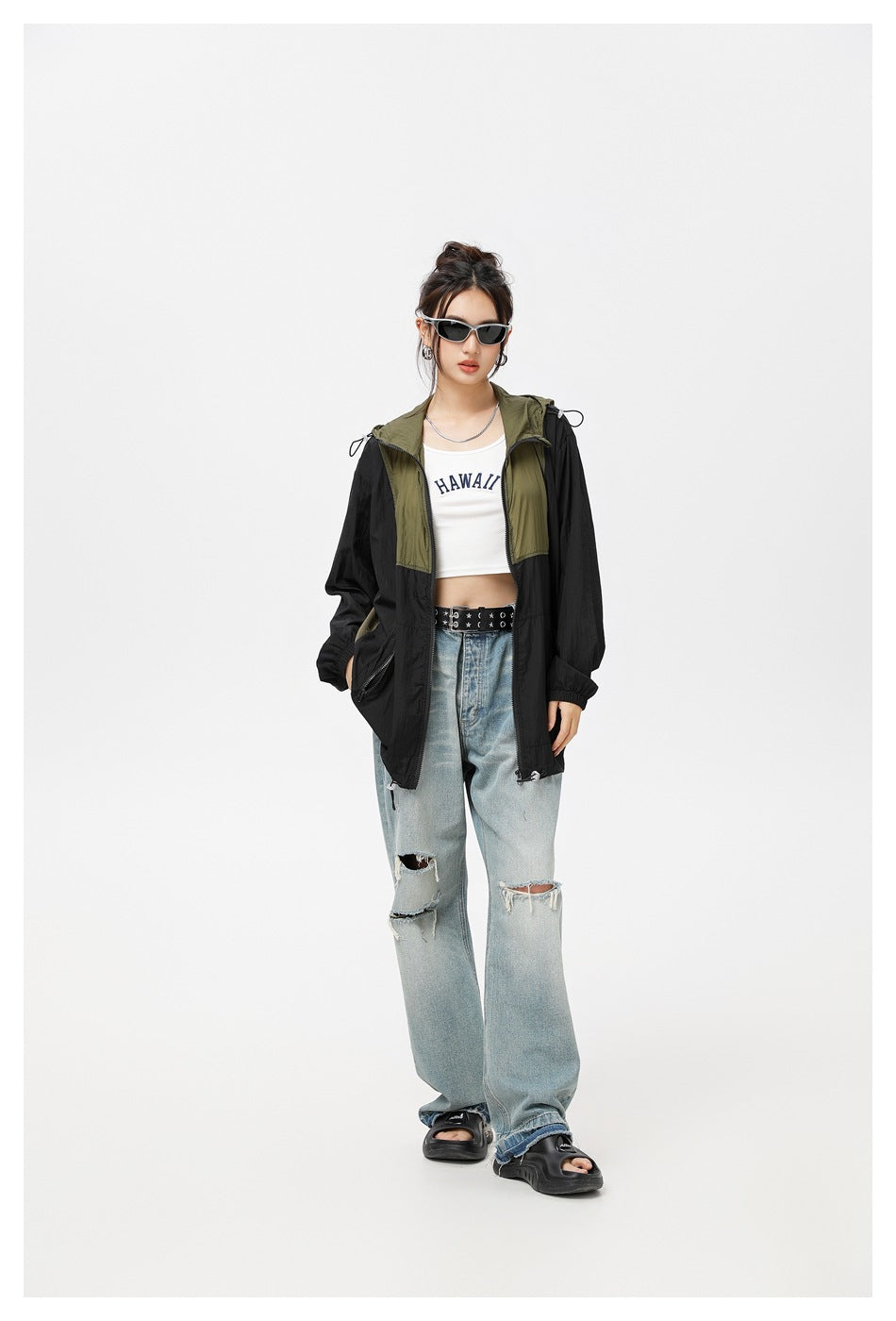 Model wearing a black hooded jacket with olive panels, open over a white crop top and light distressed jeans.