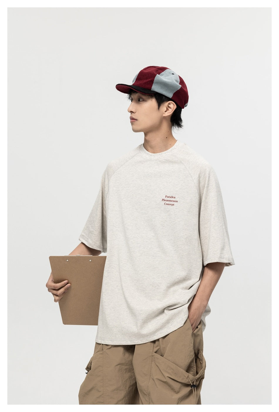 Model wearing an oversized off-white T-shirt with small burgundy chest lettering and a red-and-gray cap.
