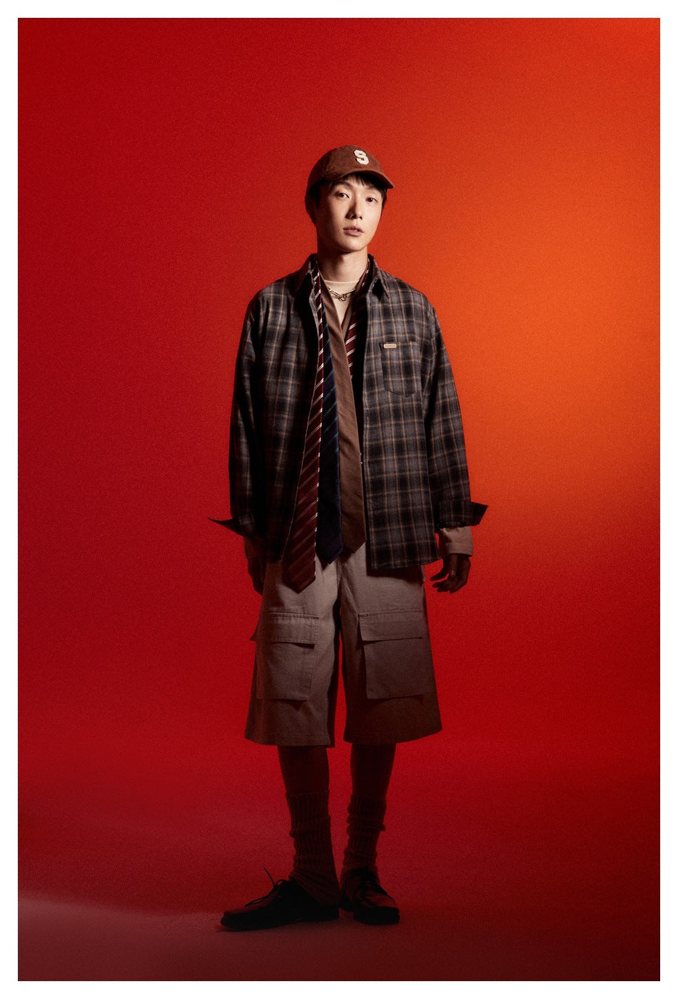 Full-length model wearing a blue-gray plaid button-up shirt with taupe cargo shorts against a red backdrop.