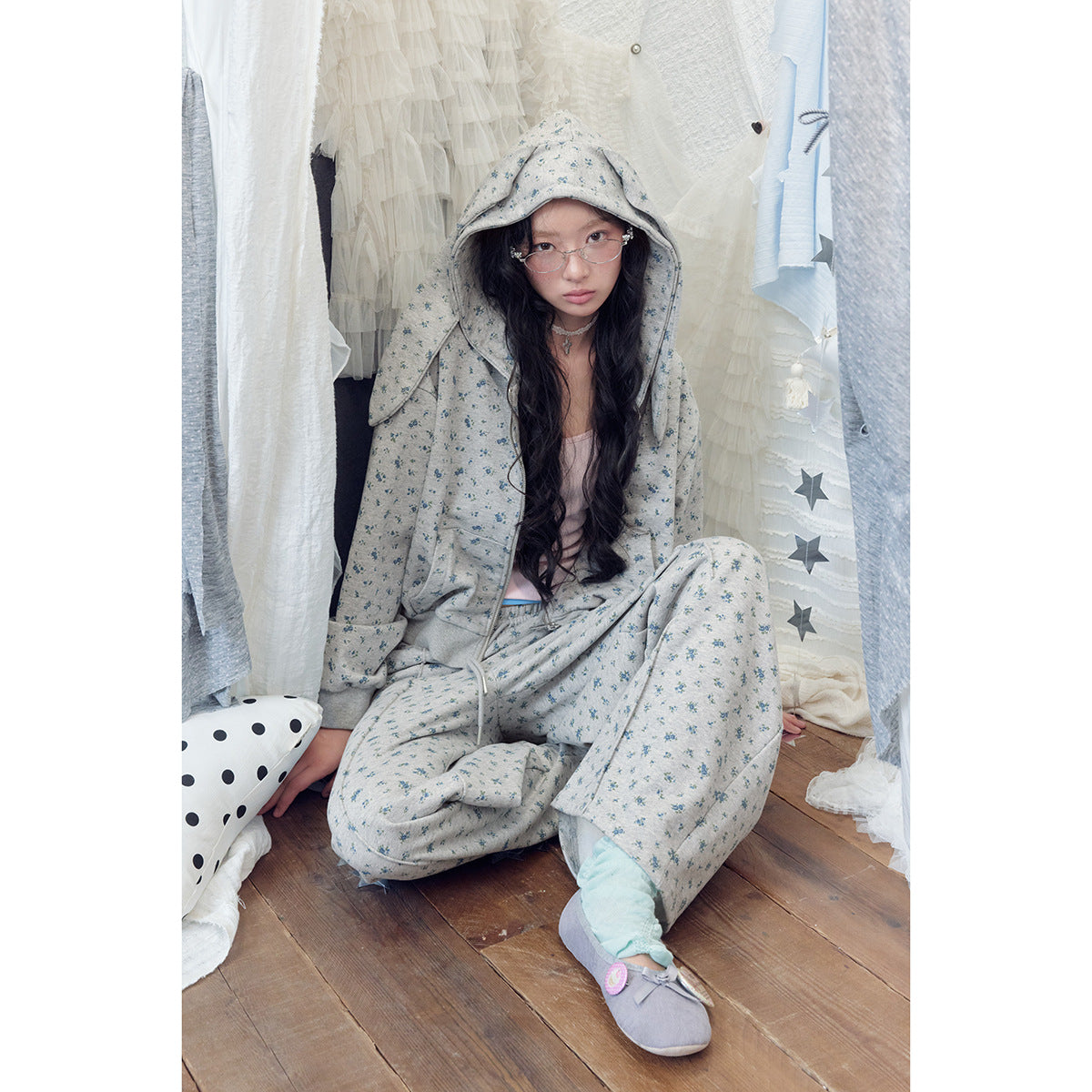 Seated model wearing a gray-blue floral zip hoodie and matching drawstring pants with the hood up.