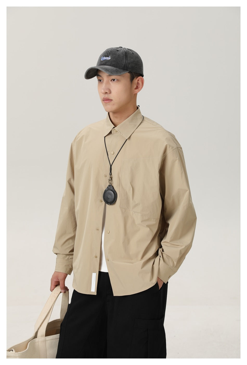 Model in a beige button-front shirt with chest pocket, carrying a tote and wearing a small black pendant.