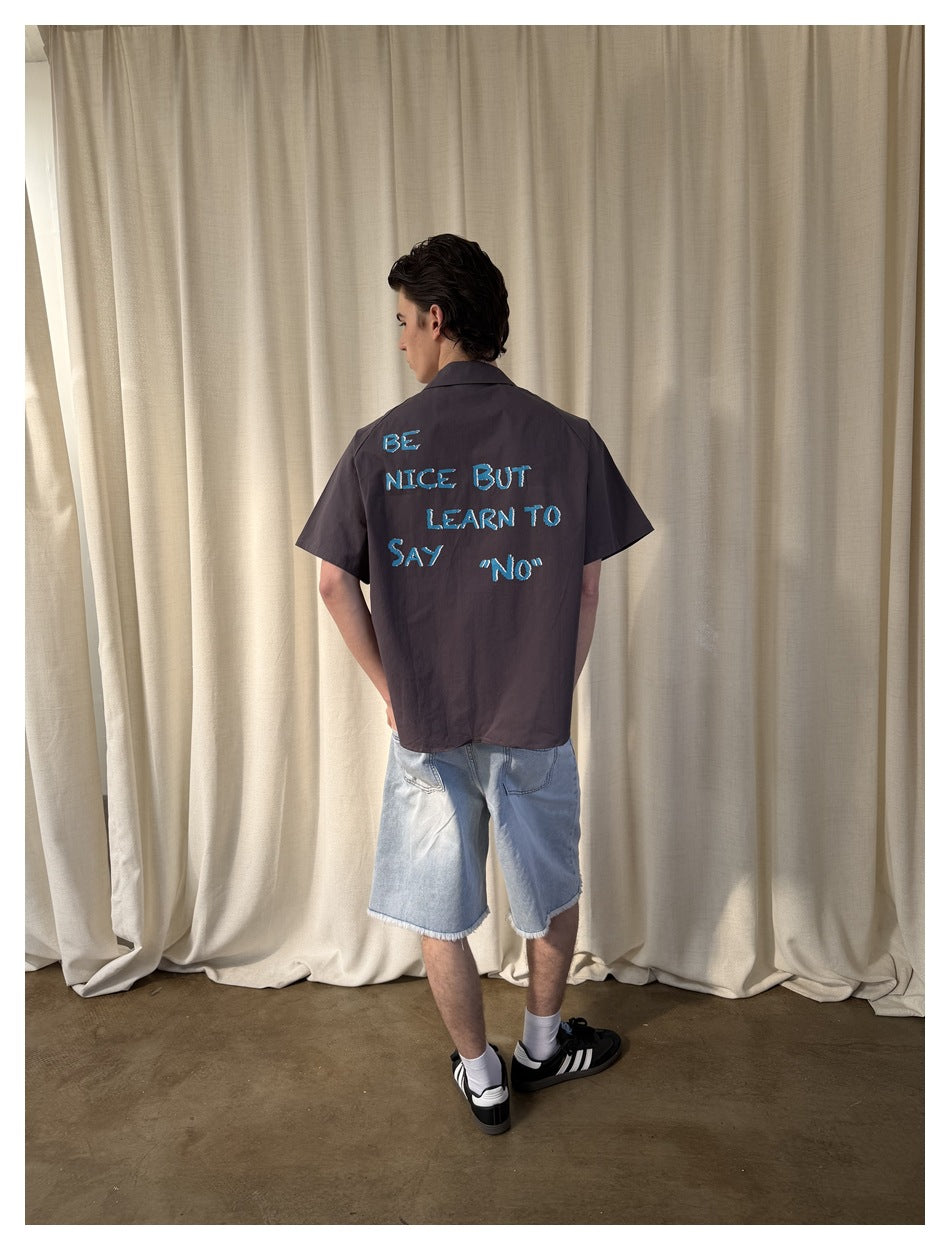 Model wearing a charcoal short-sleeve shirt with blue back lettering and light wash frayed denim shorts, rear view.