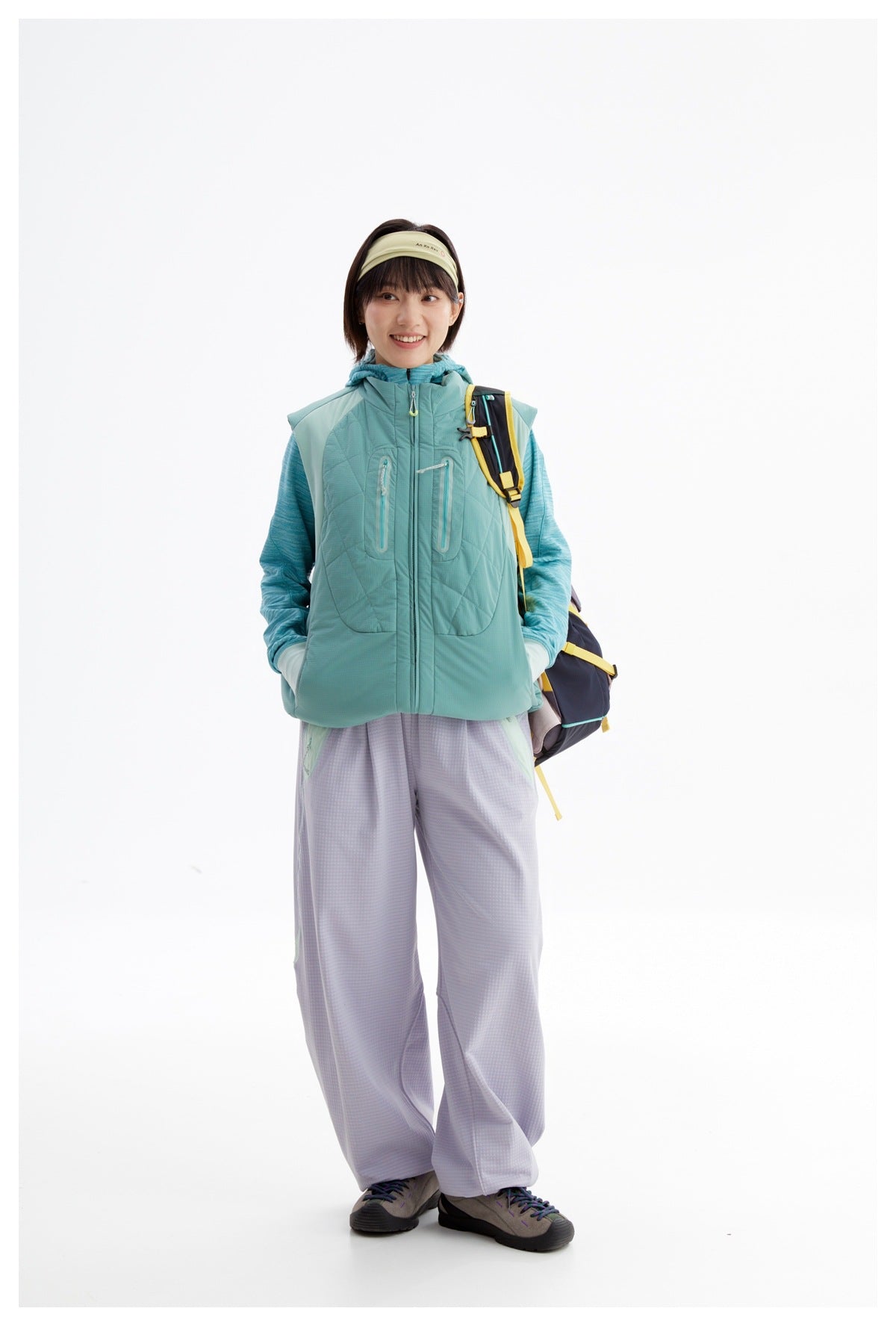 Full-length model wearing a teal quilted vest over a blue hooded top with lavender pants and a backpack.