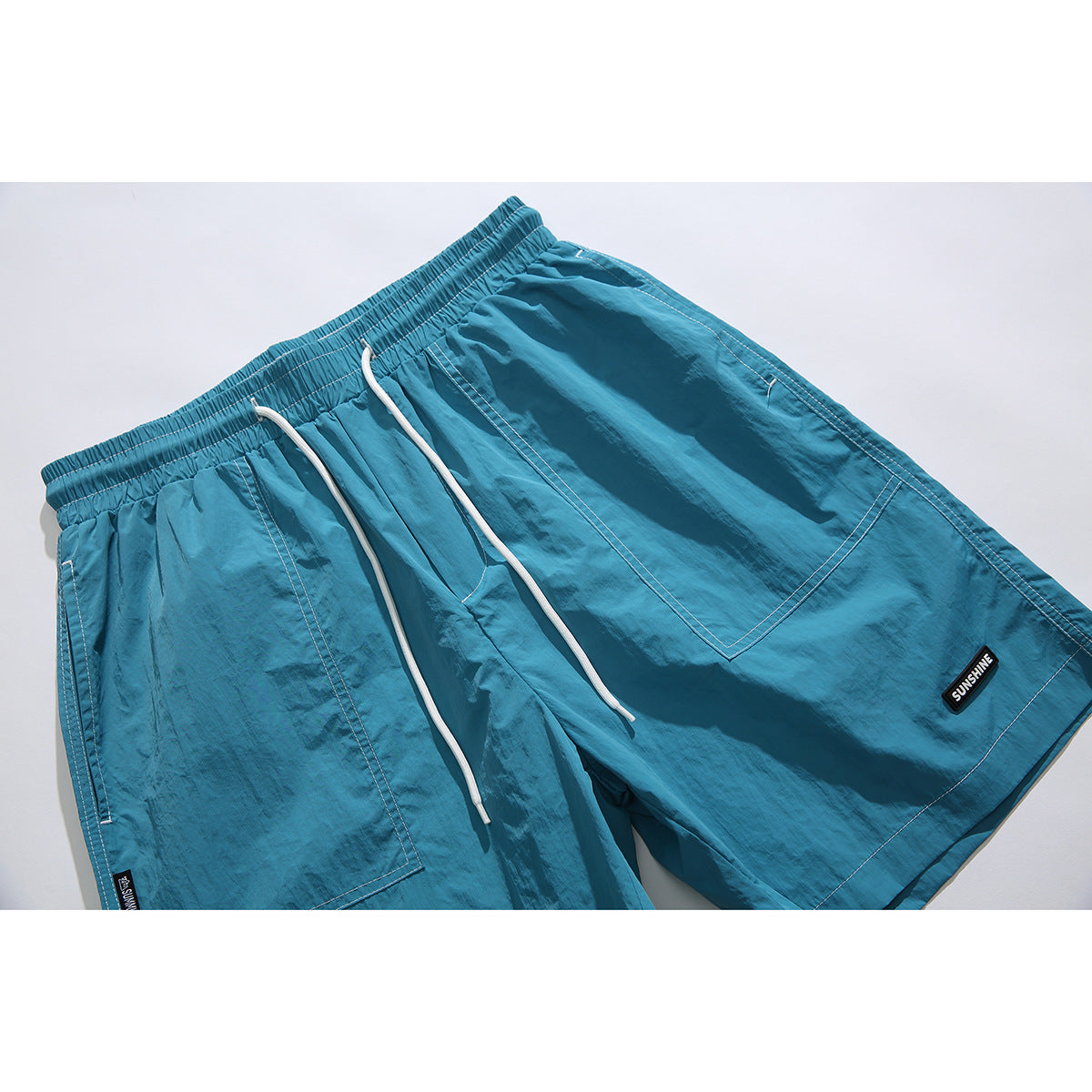 Angled flat lay of teal shorts with an elastic drawstring waist, white cords, large pockets and small black patches.