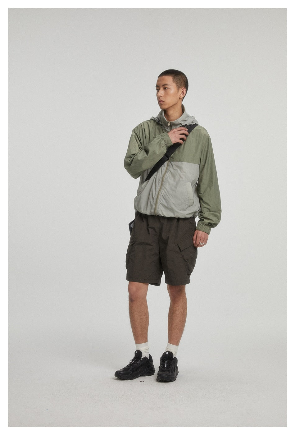 Model wearing an olive and light gray zip-front hooded jacket with dark cargo shorts and a black crossbody bag.