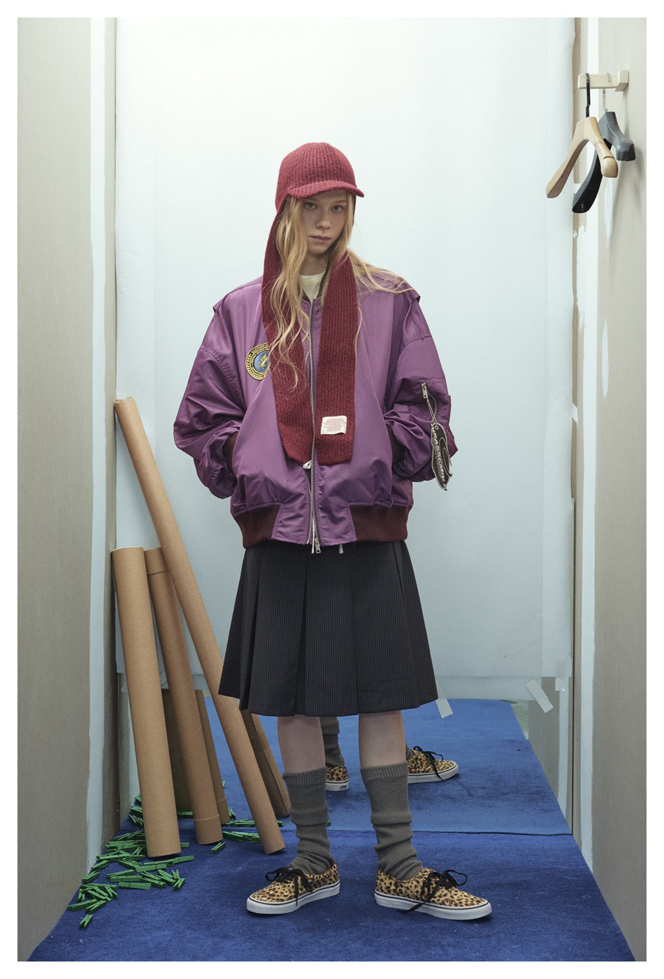 Full-length model wearing a purple bomber jacket, burgundy knit hood scarf, black pleated skirt, gray socks, and patterned