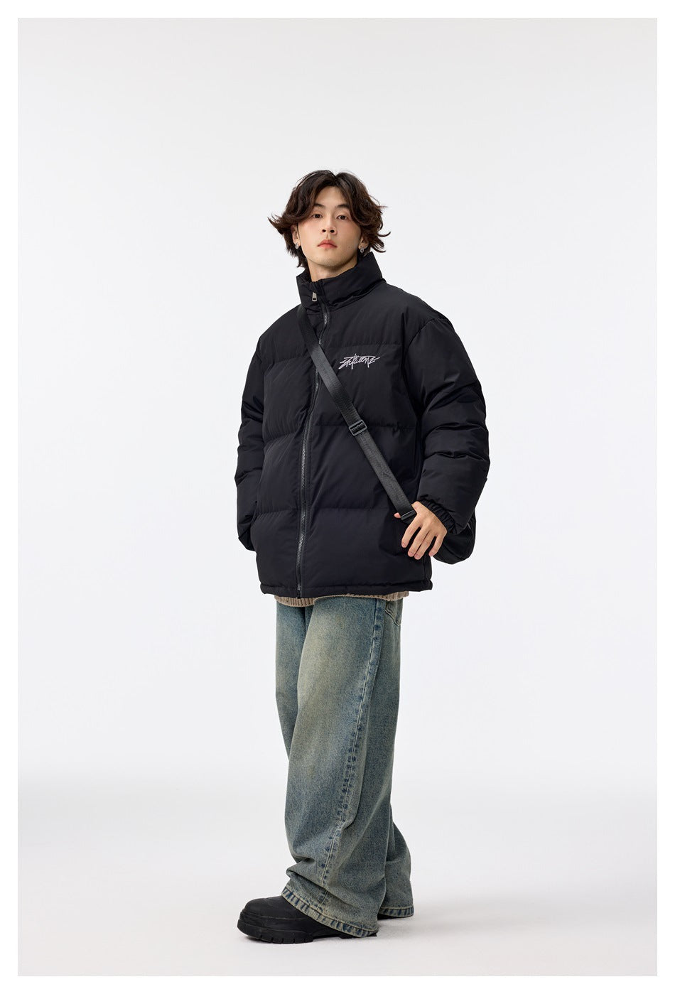 Full-length view of a model wearing a black puffer jacket with a crossbody strap and loose blue jeans.