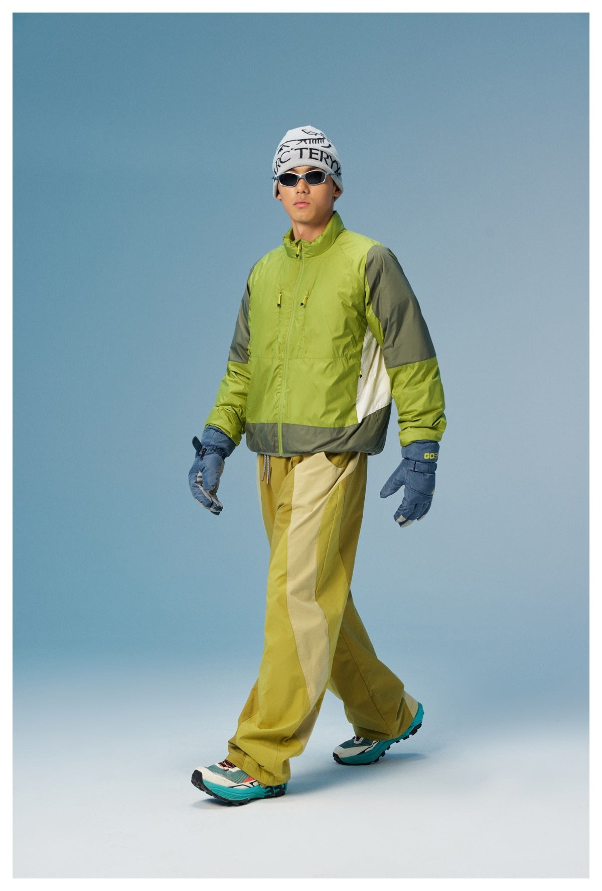 Full-length male model wearing a lime green zip jacket with gray panels and white side inserts, paired with mustard trousers.