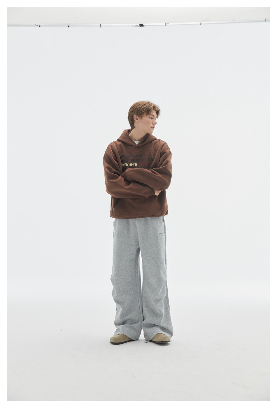 Model in a brown text-print hoodie and loose gray pants, standing with arms crossed against a white studio background.