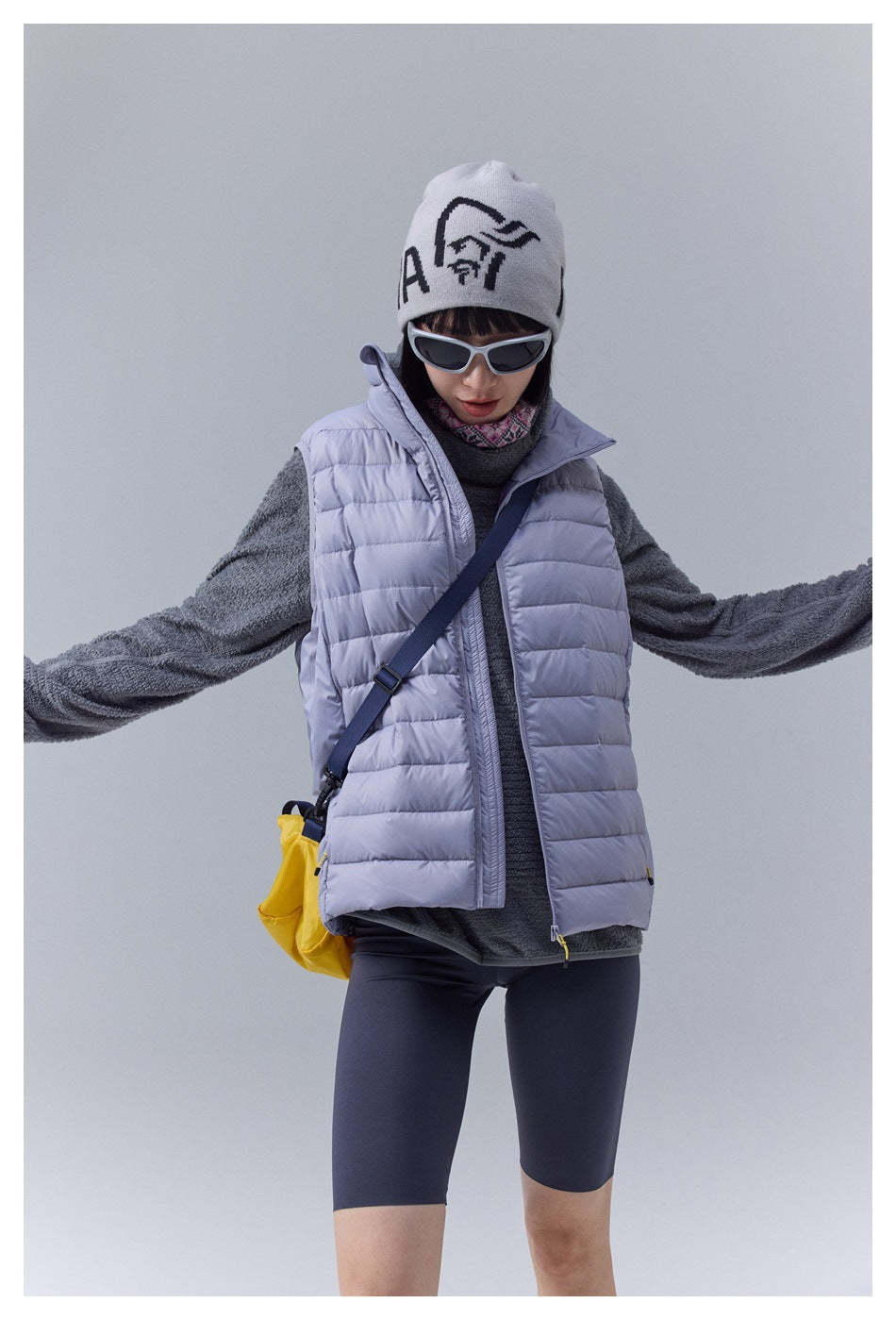 Model wearing a pale lavender quilted vest over a charcoal fleece, with dark cycling shorts and a white graphic beanie.
