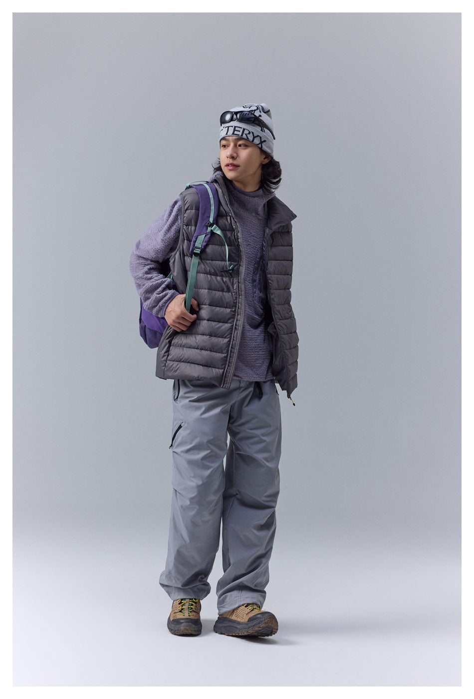 Model wearing a charcoal quilted vest over a textured purple top, with gray outdoor trousers and a backpack.