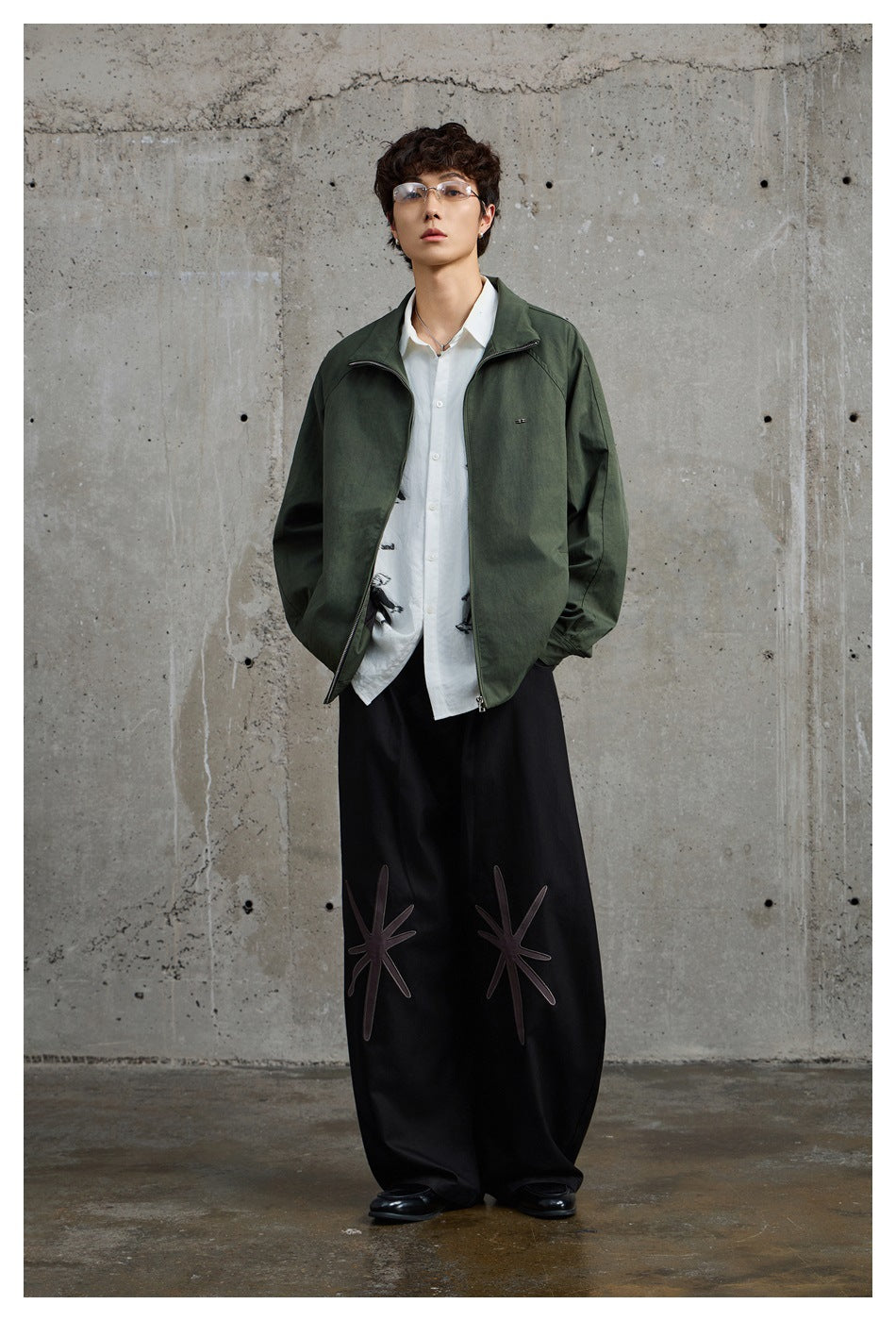 Model wearing a dark olive zip jacket over a white shirt, with wide black trousers featuring large stitched motifs.