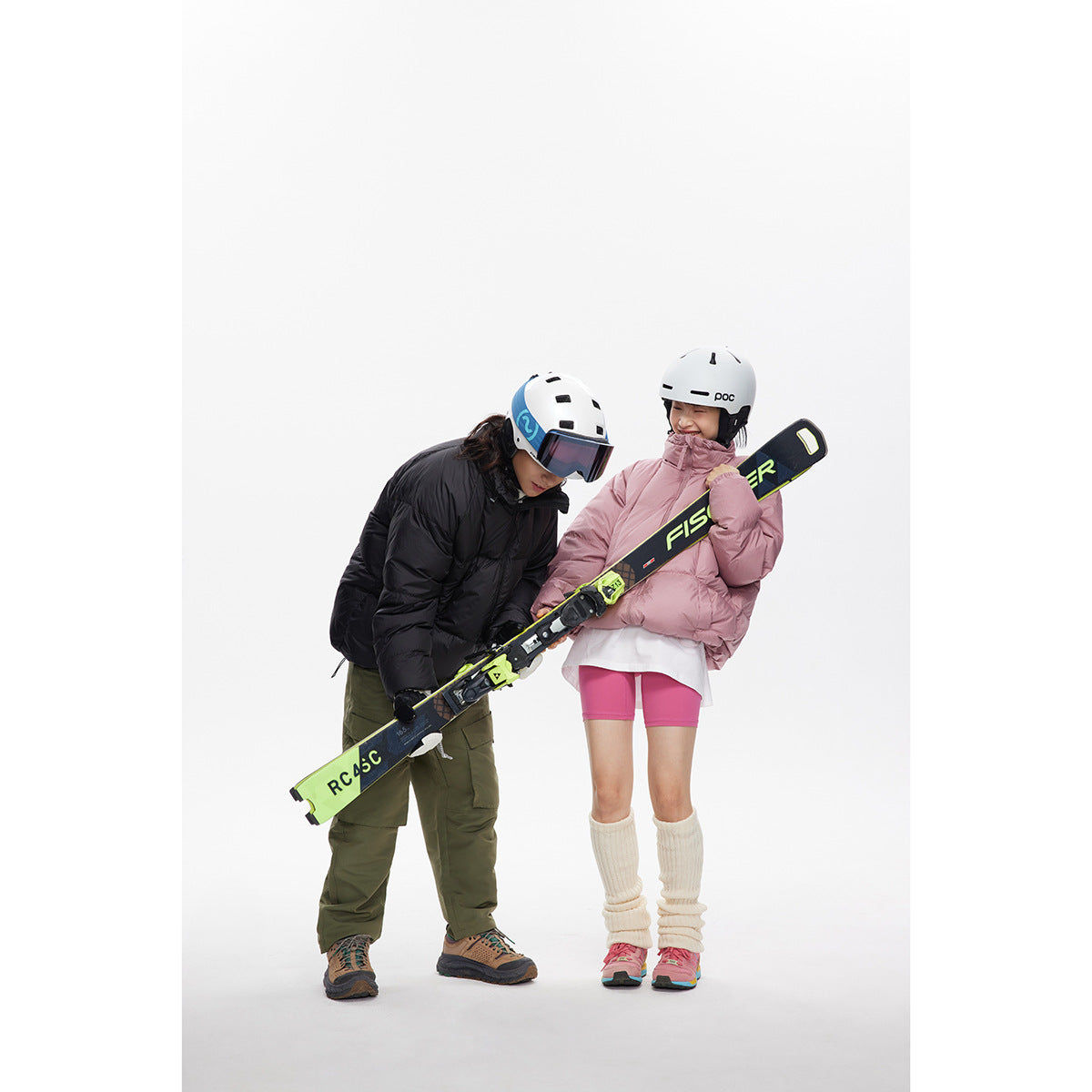 Two people in ski gear inspect a neon-accented ski; one wears a black puffer and the other a pink puffer.