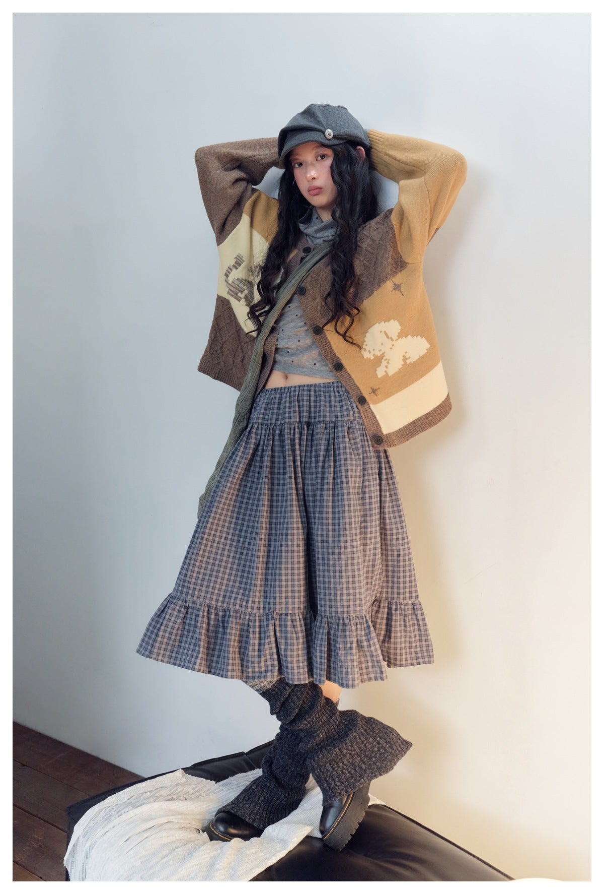 Full-length model wearing a color-block cardigan, gray top, blue plaid tiered skirt, cap and dark leg warmers.