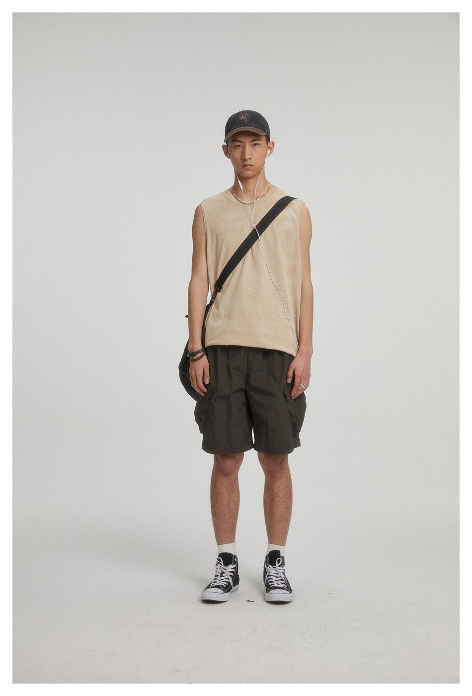 Front view of a man wearing a beige sleeveless top and dark cargo shorts, with a crossbody bag.