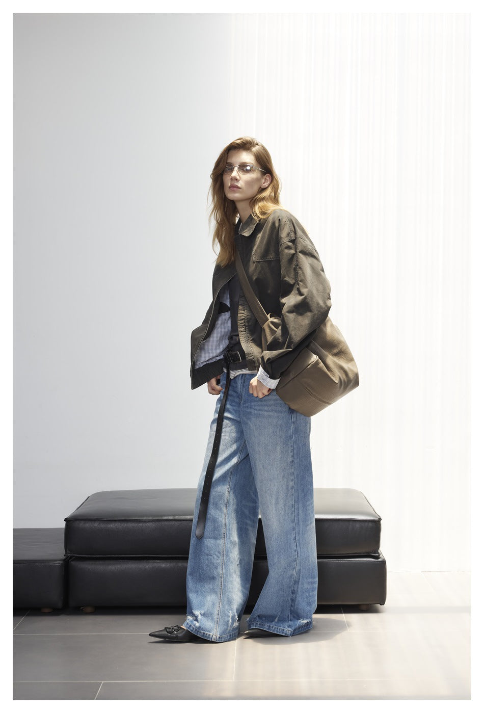 Full-length view of a model in a dark olive oversized jacket and wide-leg blue jeans, carrying a taupe shoulder bag.