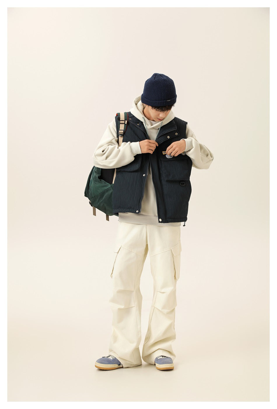 Model wearing a dark charcoal quilted utility vest with flap pockets over an ivory hoodie and cargo pants.